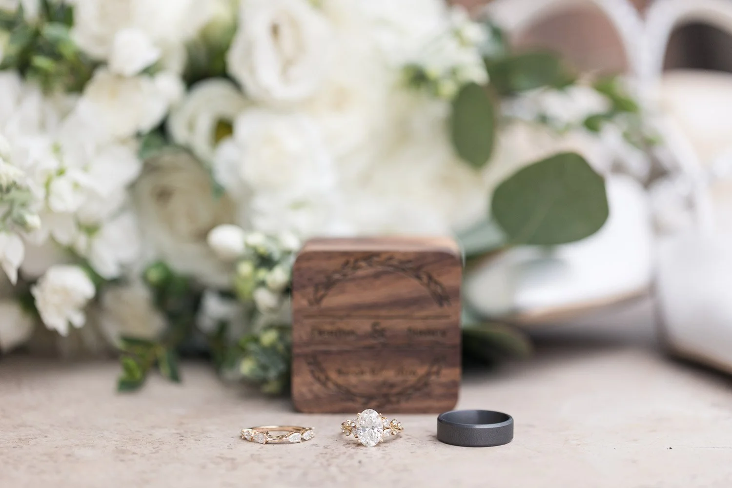 Wedding rings and engagement ring in front of a bouquet of white flowers and greenery, with a wooden box in the background.