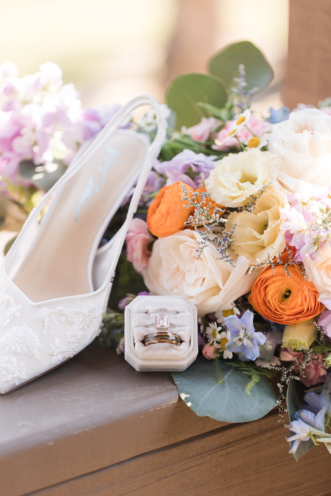 A silk white bridal shoe with intricate lace embroidery, a small white jewelry box withdiamond rings, and a colorful bouquet of roses and other flowers on a wooden surface.