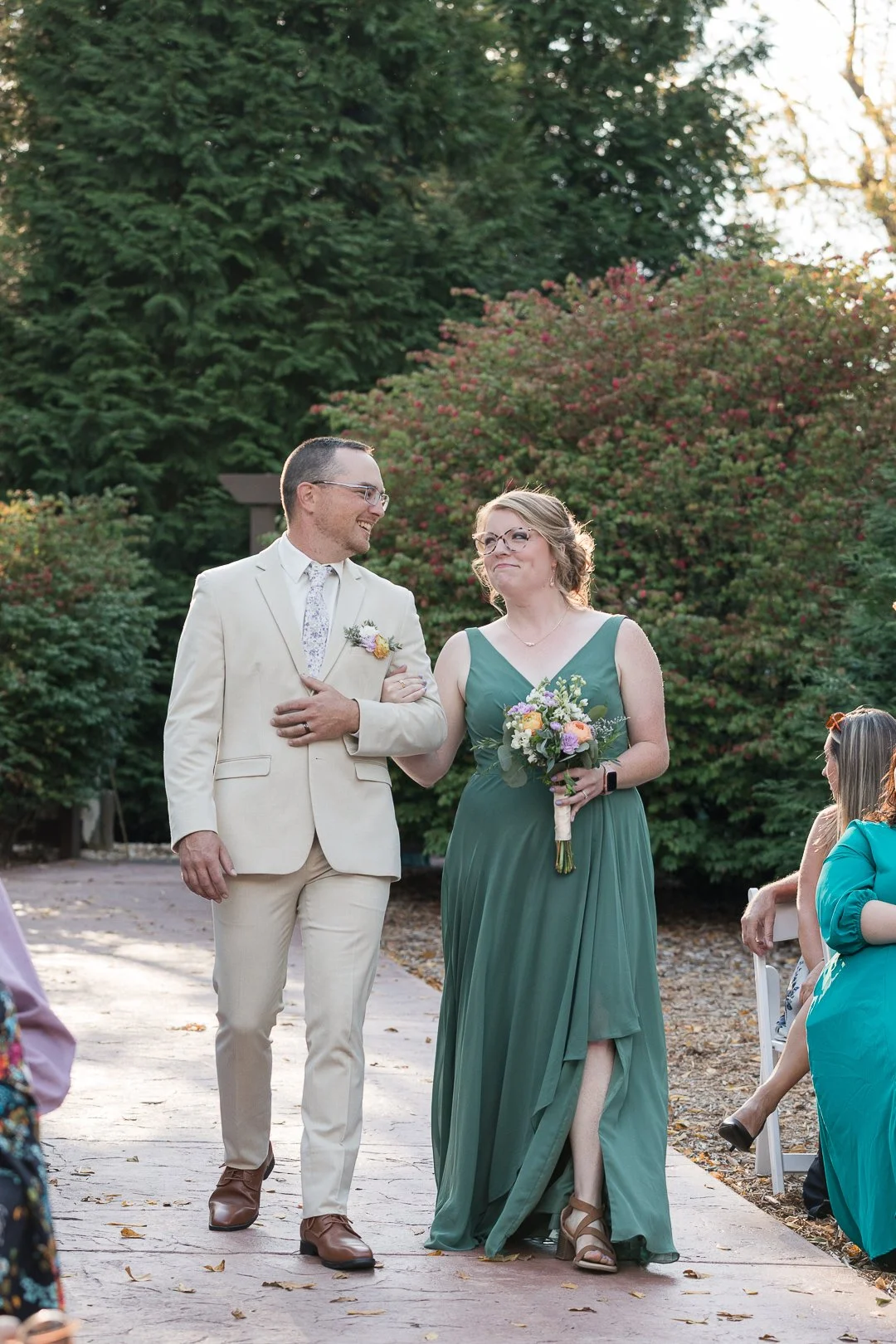 A groom and a woman, possibly a bride or bridesmaid, walking arm in arm outdoors during a wedding ceremony, surrounded by greenery and seated guests.