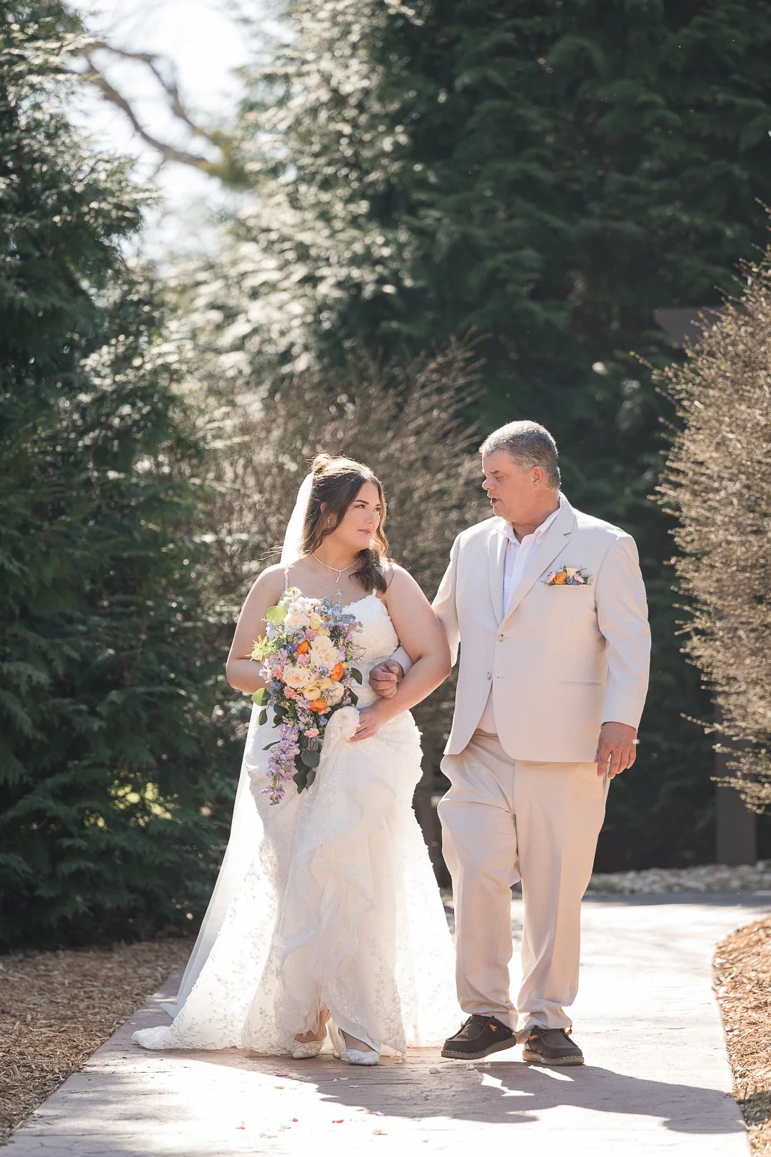 A bride in a white wedding gown holding a colorful bouquet, walking arm-in-arm with an older man in a cream suit on a garden pathway surrounded by trees.