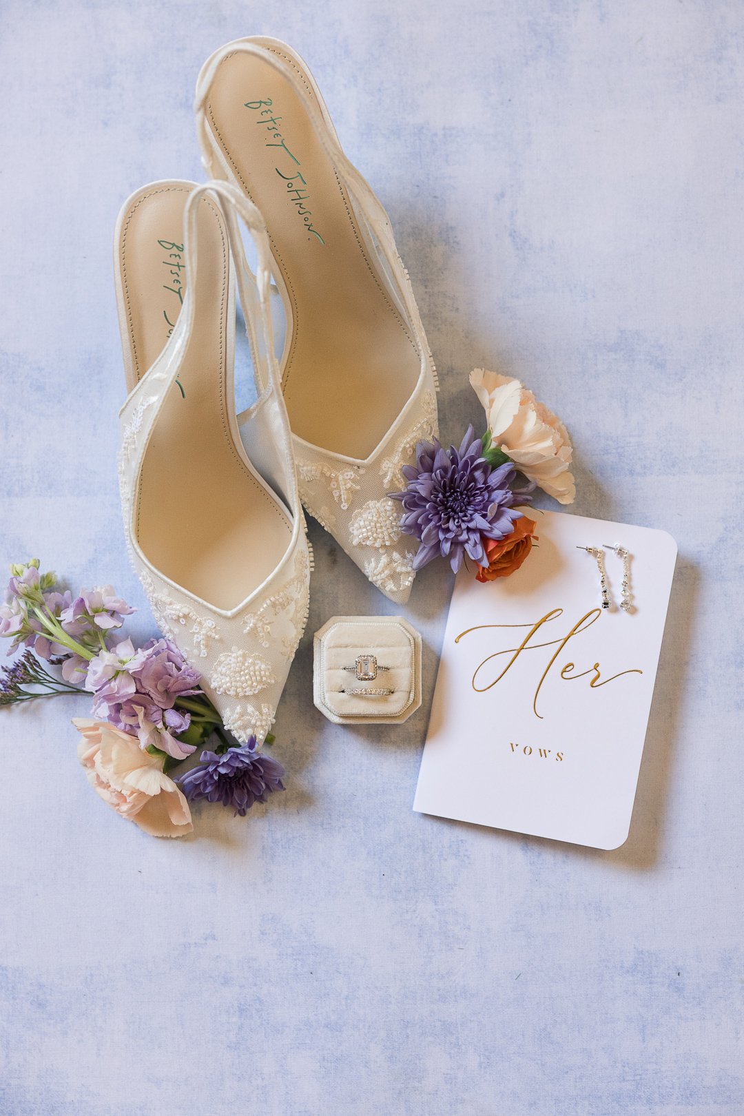 White bridal shoes with floral embellishments, a pair of earrings, a ring in a velvet box, and a 'Her' card surrounded by pastel flowers.