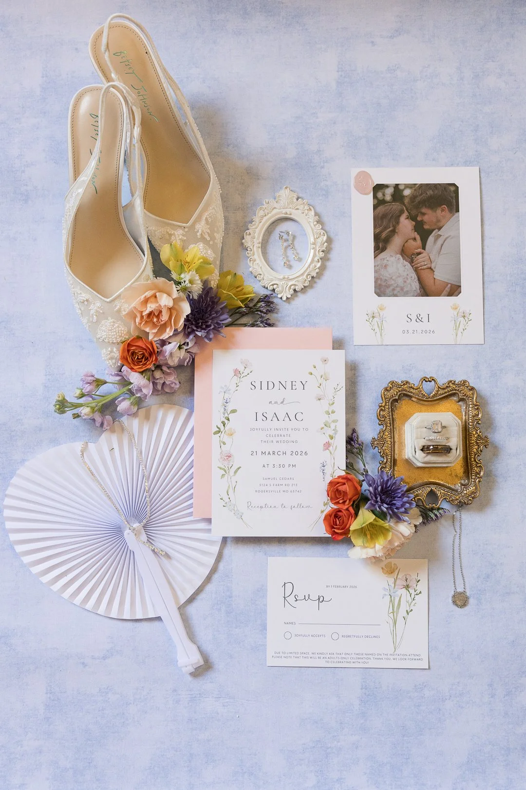 Wedding flat lay with ivory heels, floral bouquet, invitation card, RSVP card, photo of a couple, rings in a decorative box, pearl necklace, photo frame, and decorative fan arranged on a light surface.