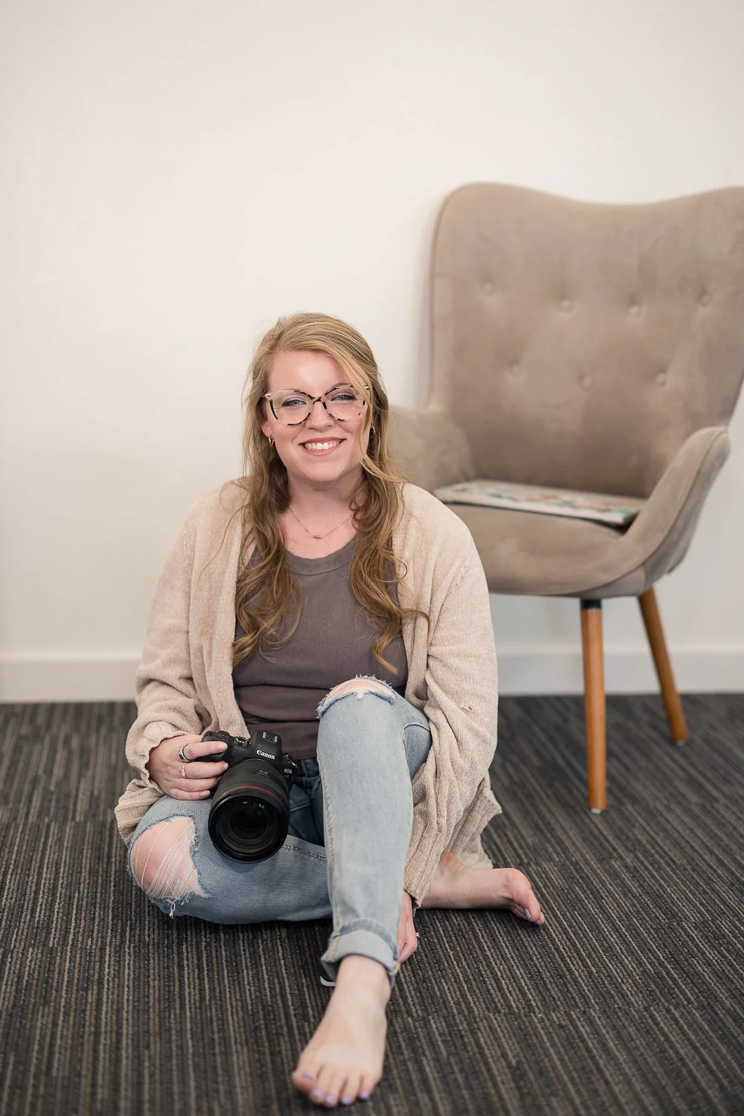 A smiling woman with glasses and long, wavy hair sitting on a carpeted floor, holding a camera, in front of a white wall and a modern beige armchair.