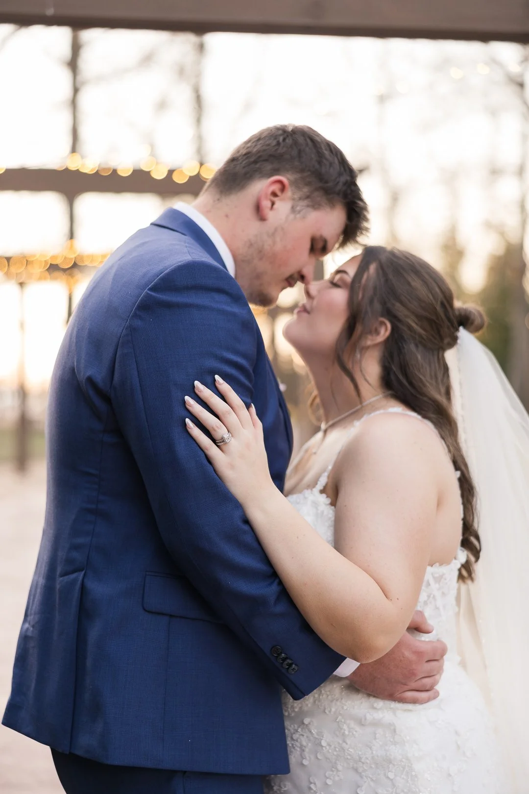 A newlywed couple embracing and touching foreheads outdoors during sunset, with blurred string lights in the background.