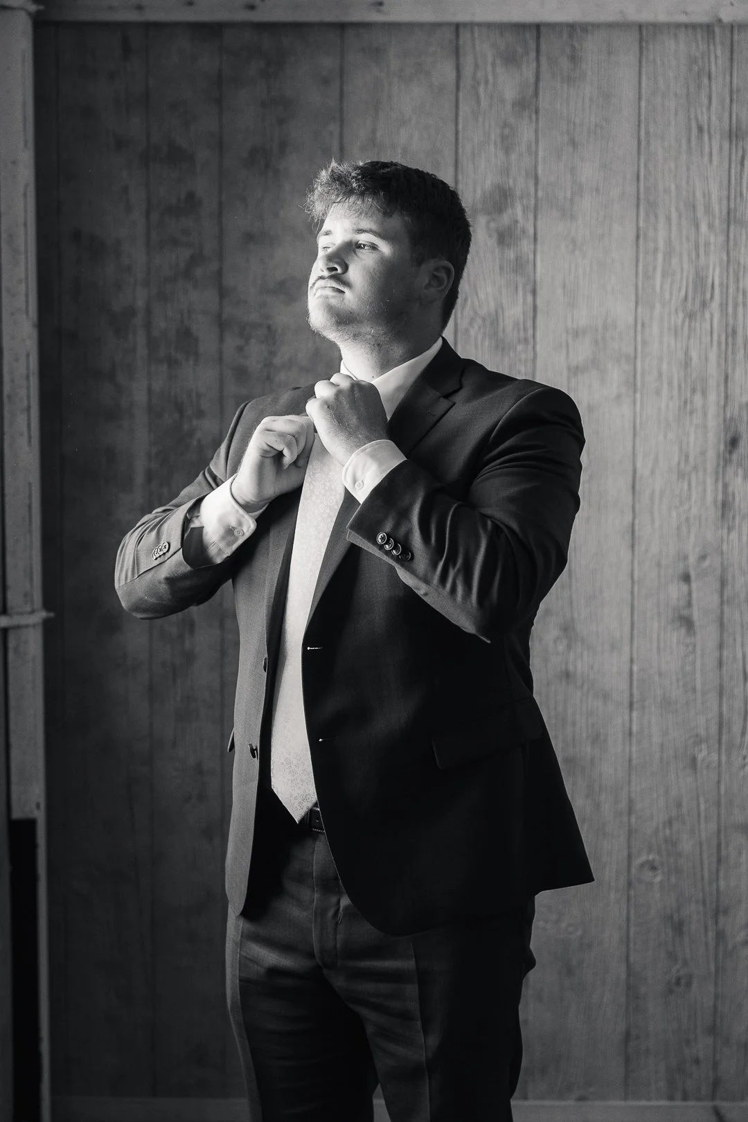 A young man in a suit adjusting his tie, standing in front of a wooden wall, black and white photograph.