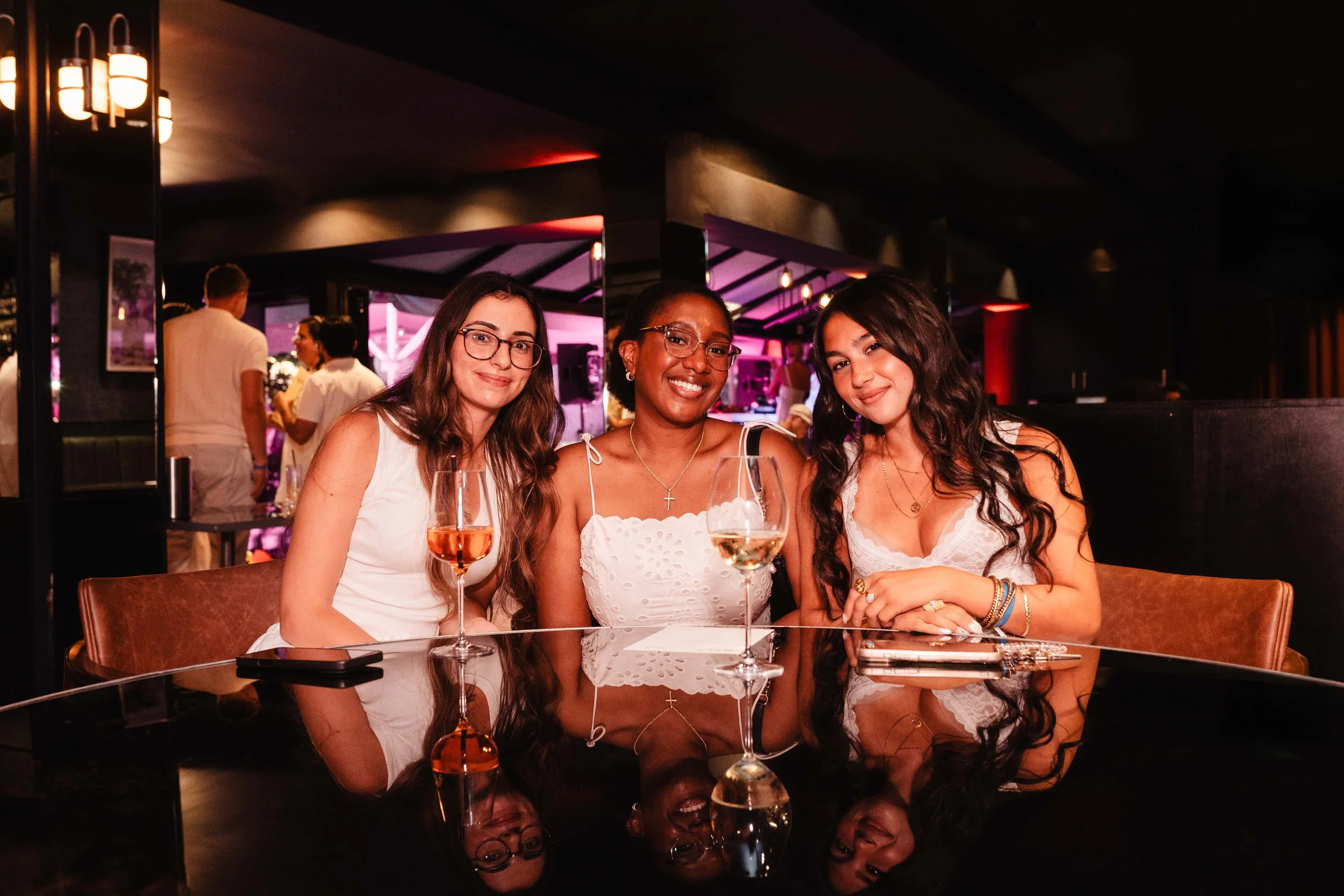 Three women sitting together at a restaurant table, smiling, with wine glasses in front of them and a dark, stylish interior background.