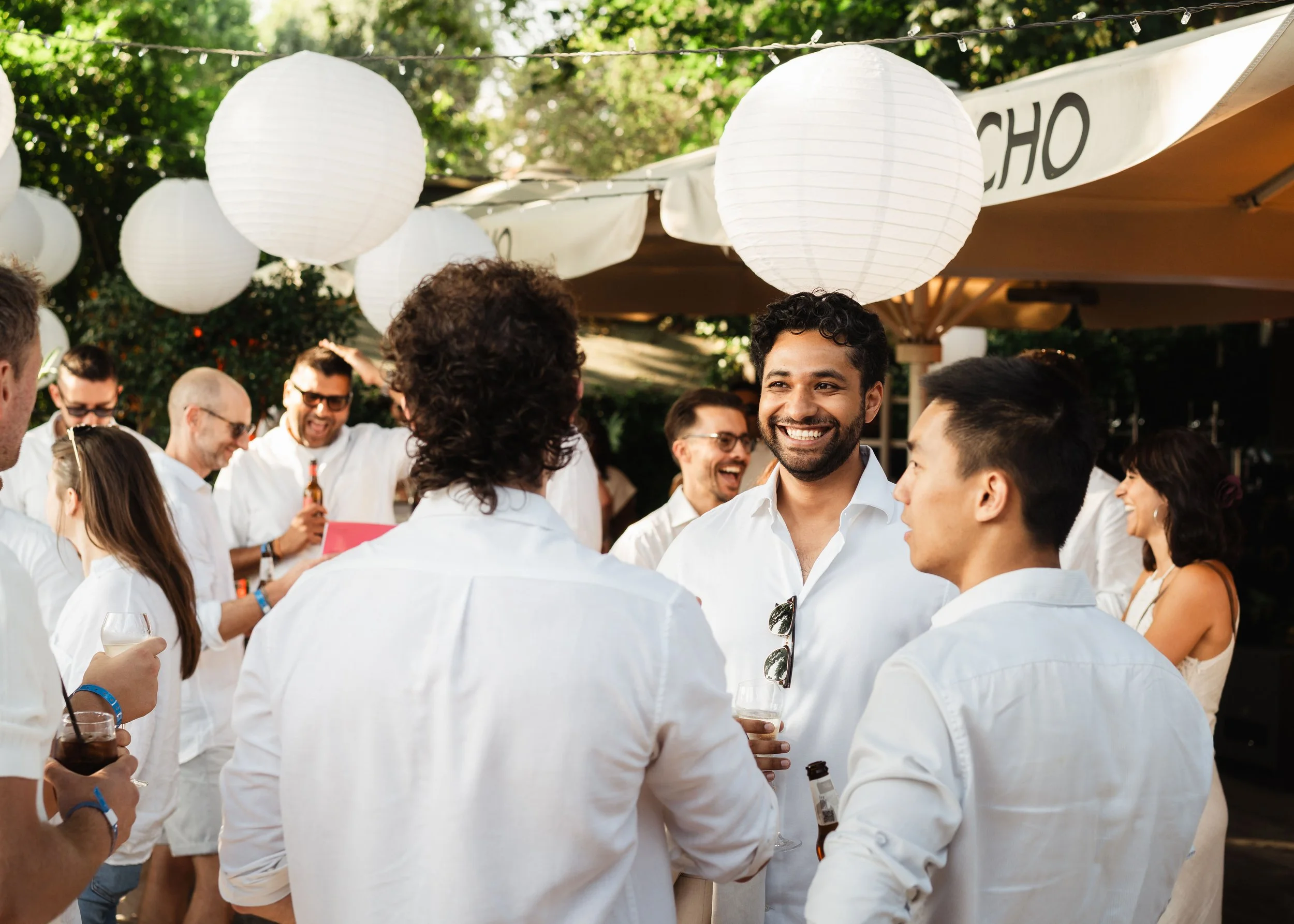 Group of people at an outdoor gathering, wearing white shirts, under hanging white paper lanterns, smiling and engaging with each other.
