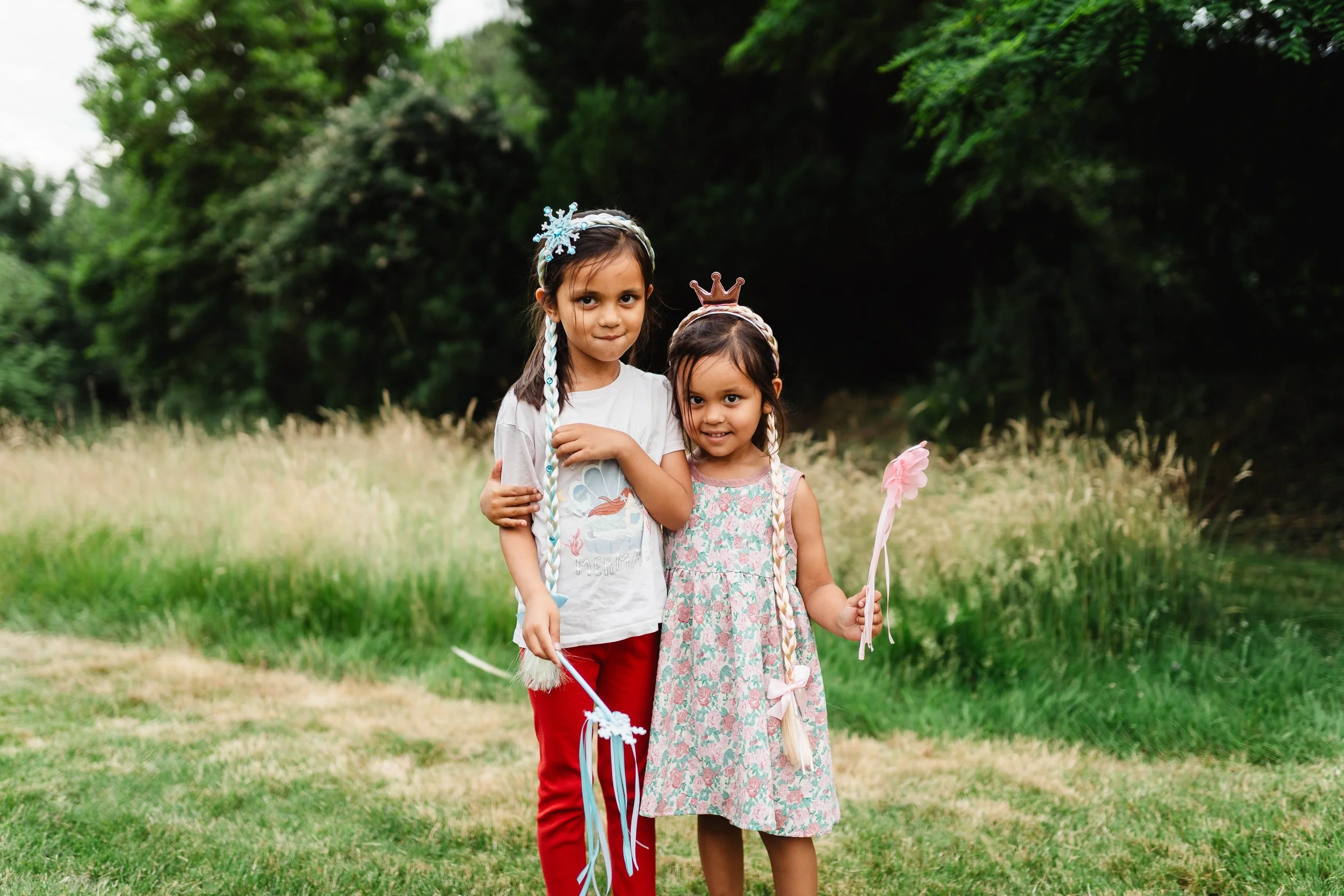 Two young girls standing outdoors, holding fairy tale wands, with one wearing a princess crown. They are in a grassy area with green trees in the background.