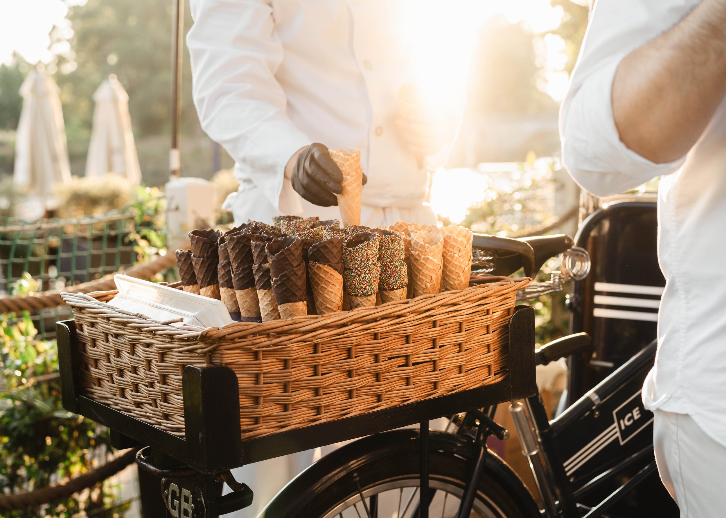 A person pushing a bicycle with a basket filled with ice cream cones, some decorated with chocolate and colorful sprinkles, outdoors during sunlight.