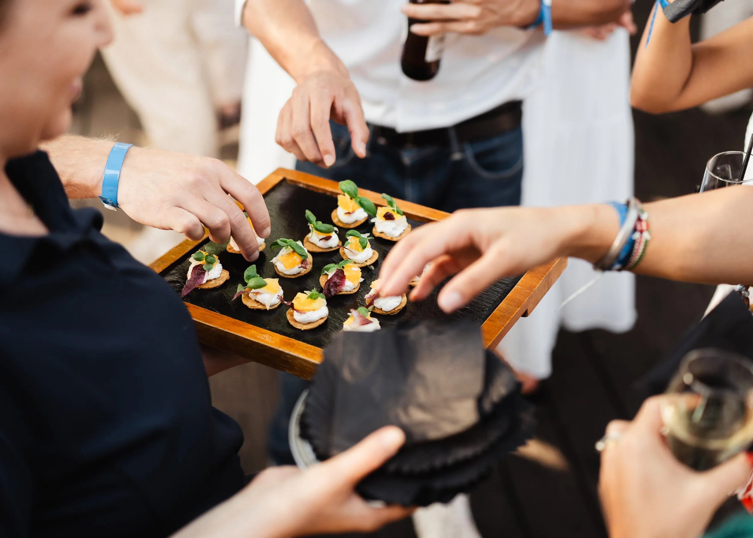 People reaching for small appetizers on a wooden tray at a social gathering.
