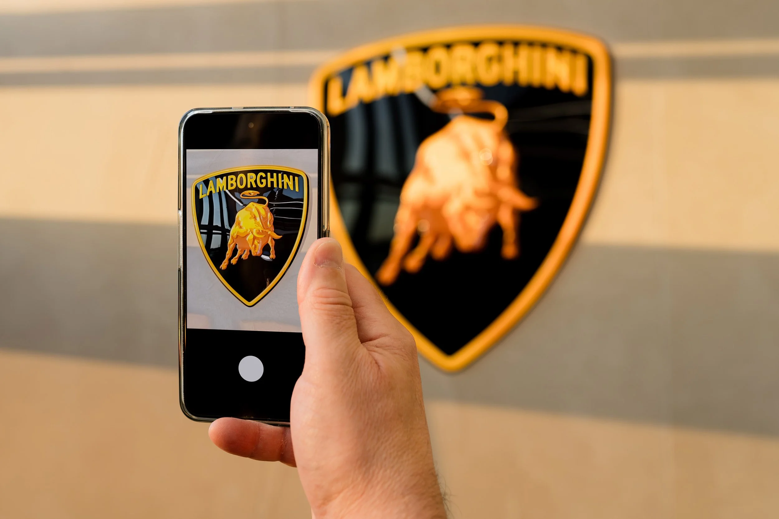 Person taking a photo of a Lamborghini logo with a smartphone in front of a Lamborghini crest on a wall.
