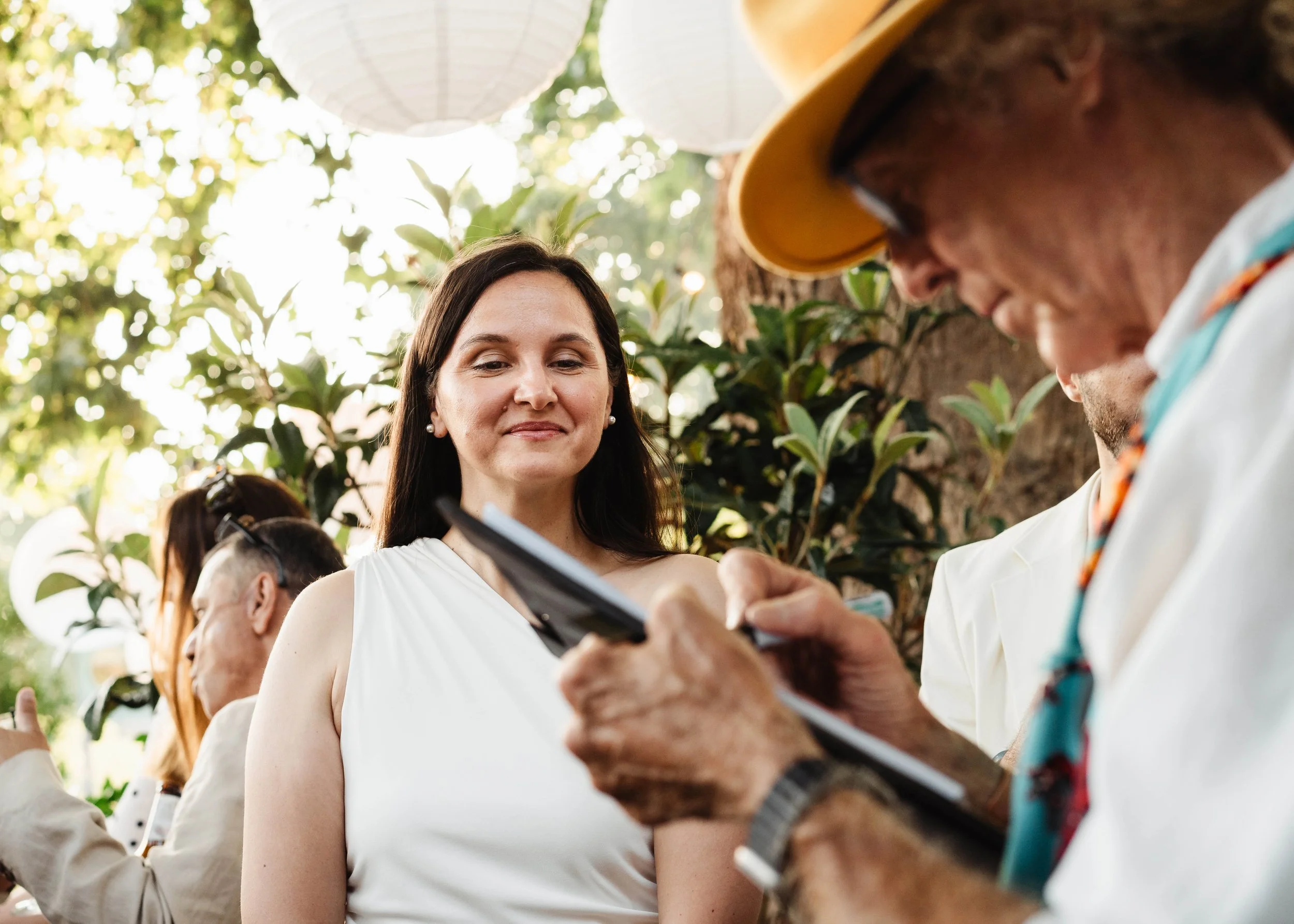 A woman with long dark hair, wearing a white sleeveless dress, smiling and looking at a man with curly hair and glasses, who is holding a phone or a device, in an outdoor setting with green trees and paper lanterns hanging above.
