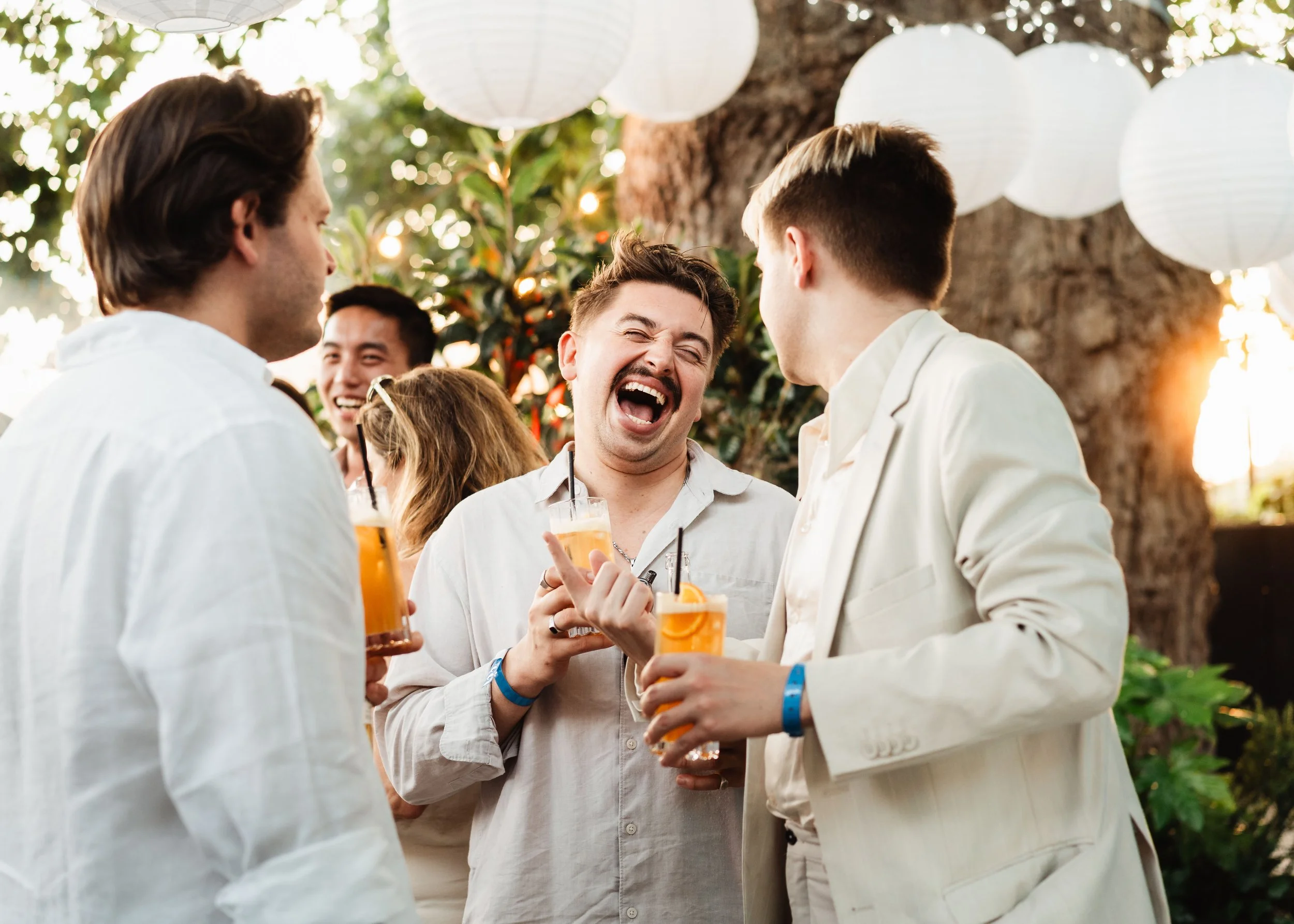 A group of young adults socializing outdoors, holding orange drinks with straws, in front of a background of hanging white paper lanterns and green foliage.
