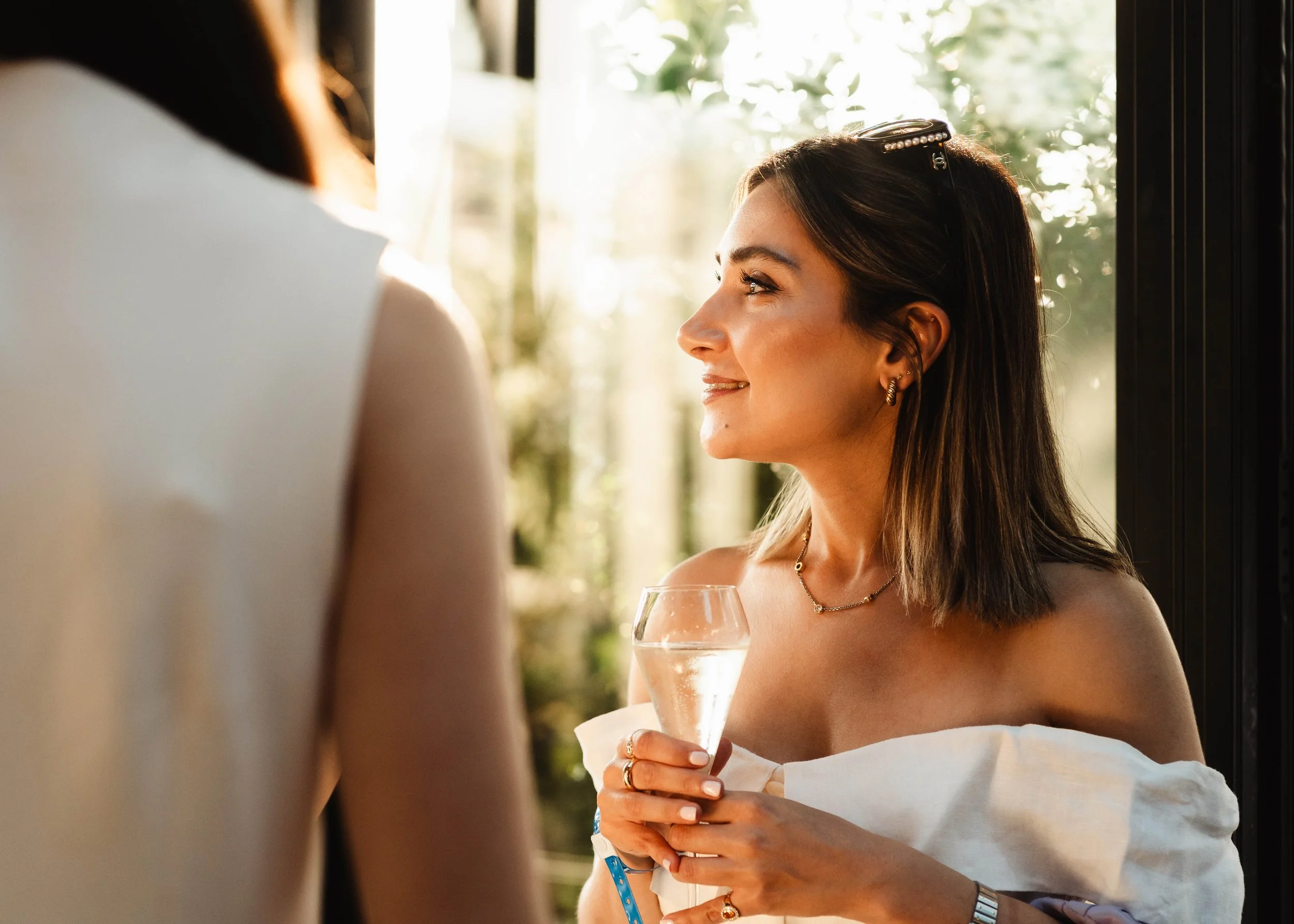 A woman with dark brown hair, wearing jewelry, holding a glass of white wine, smiling and looking at another person at a social gathering in a well-lit indoor space with large windows and greenery outside.