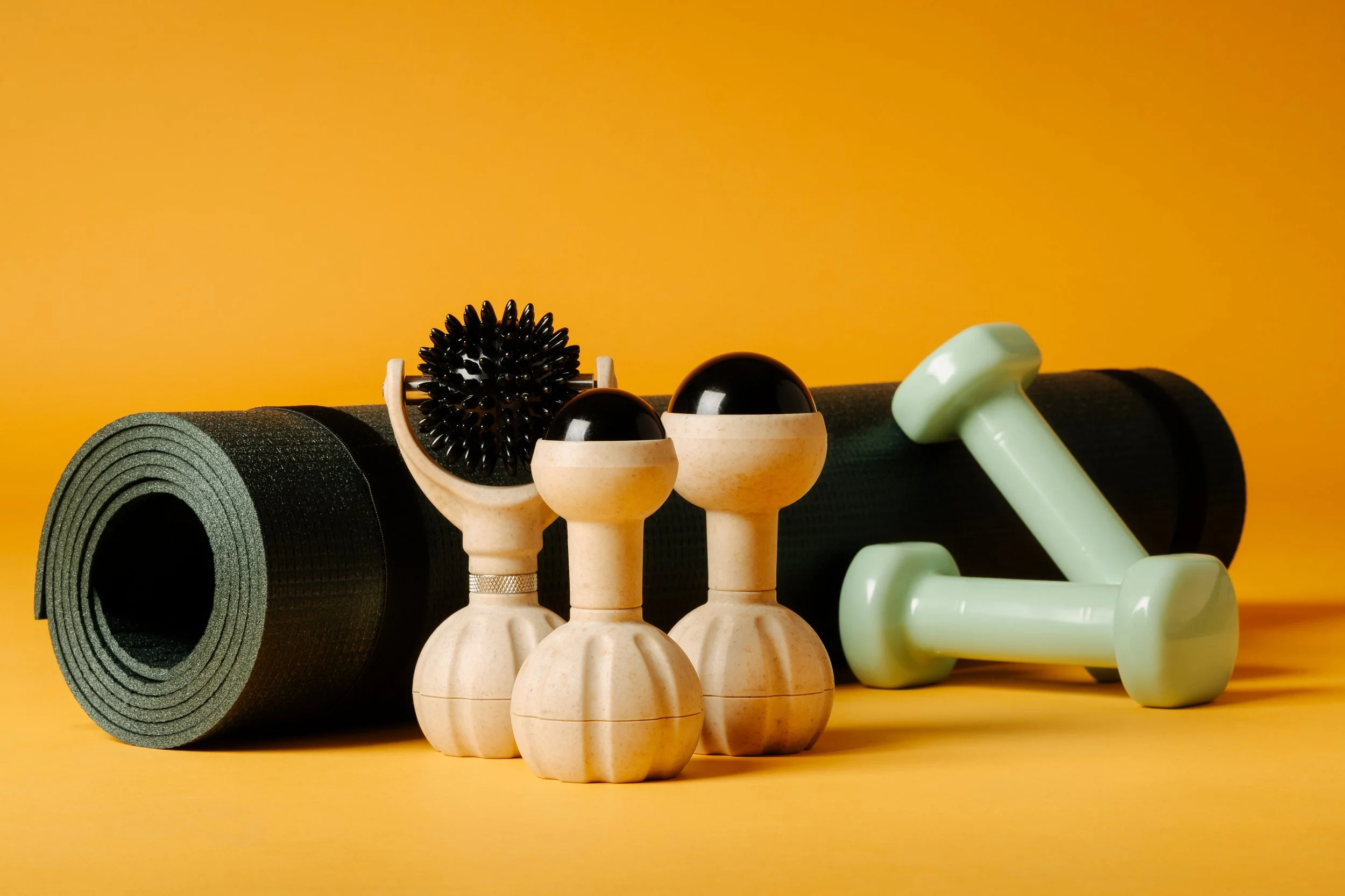 A black yoga mat roll, a massage roller with textured black surface, two beige massage cups, a light green dumbbell, and a light green massage stick arranged against a yellow background.