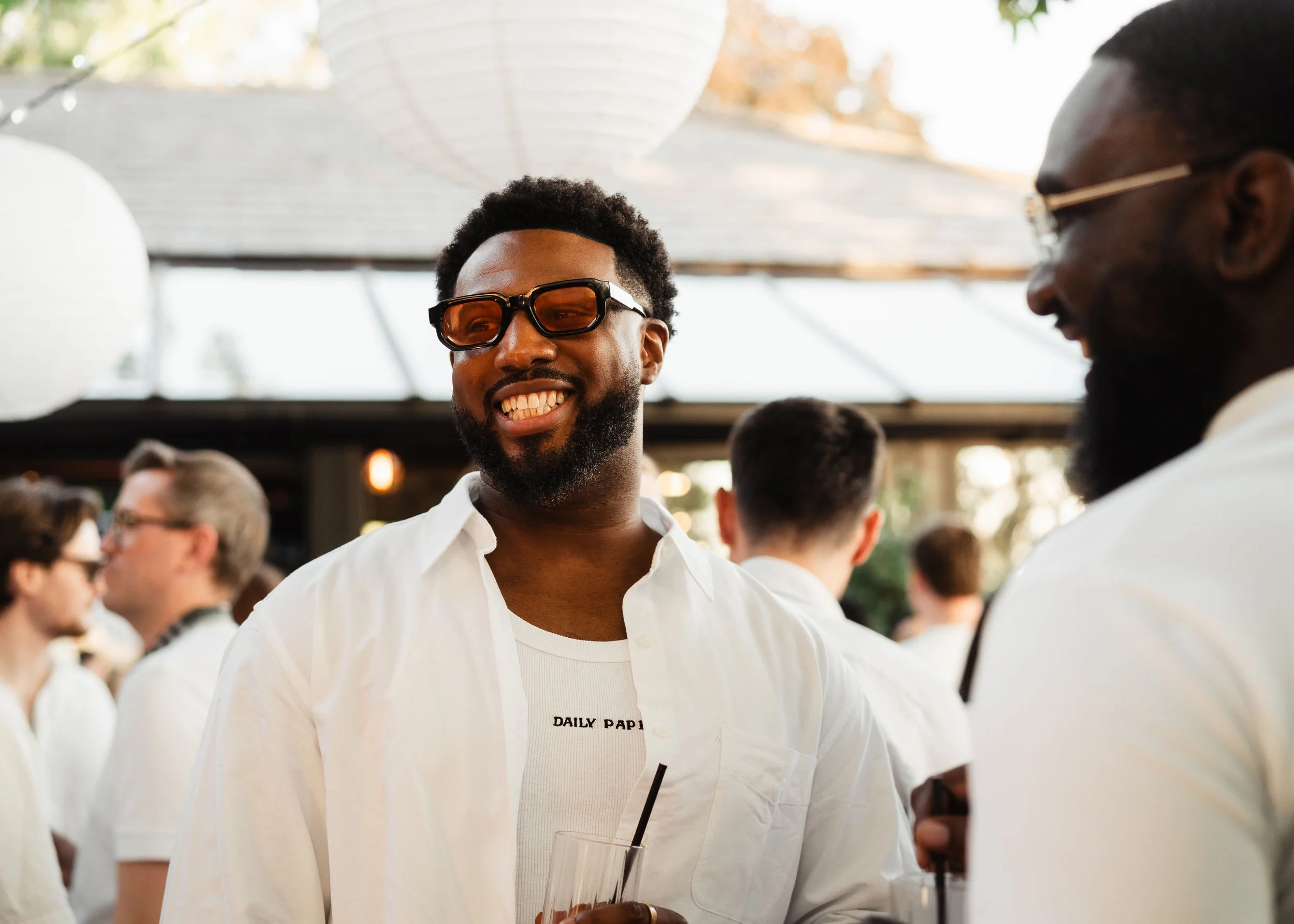 Two men with dark skin wearing white shirts and sunglasses are talking at an outdoor gathering. The man in the foreground has white text on his shirt that says "DAILY PAP," and the man in the background has a beard and is smiling.