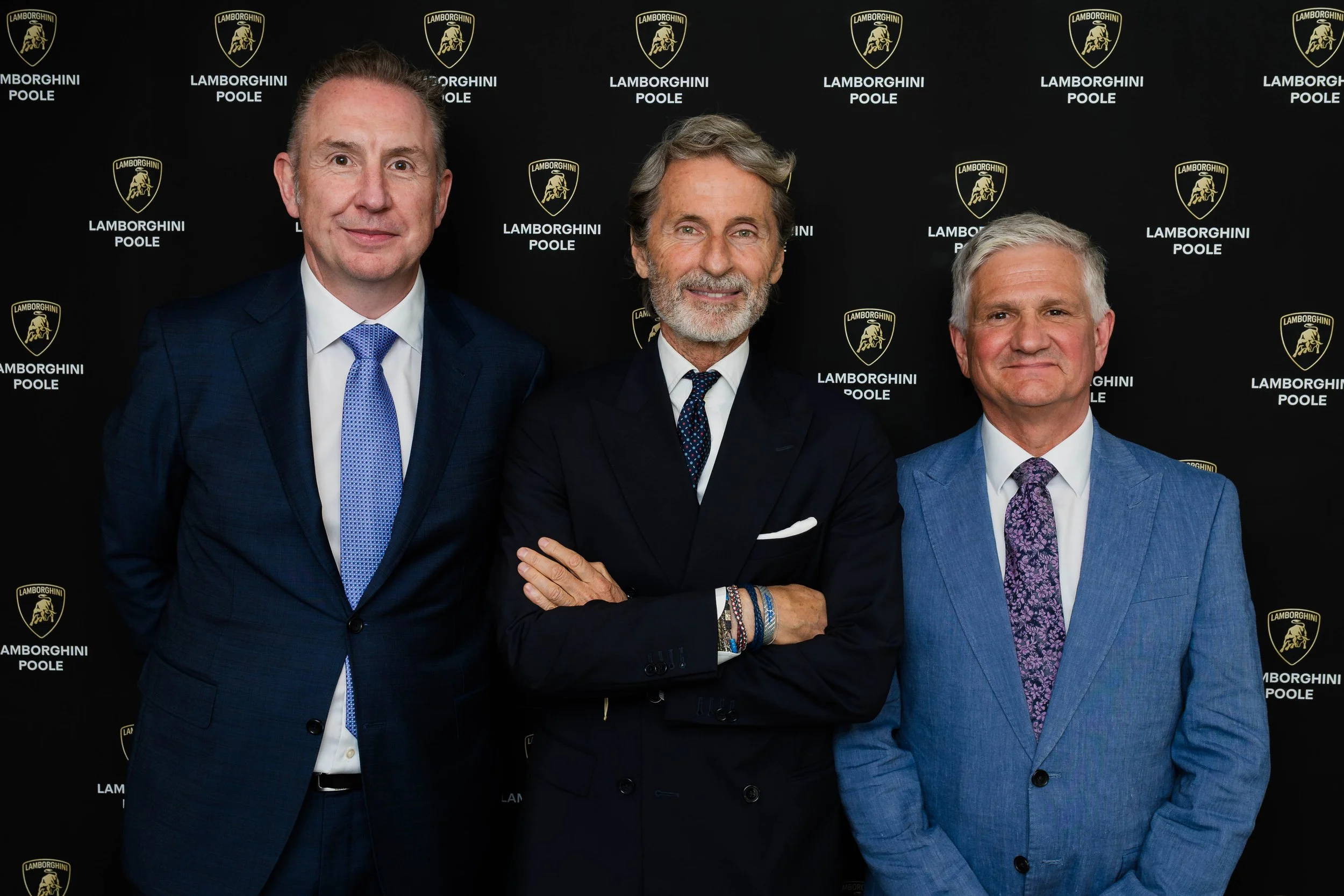 Three men in suits standing in front of a black background with Lamborghini and Poole logos, smiling at the camera.