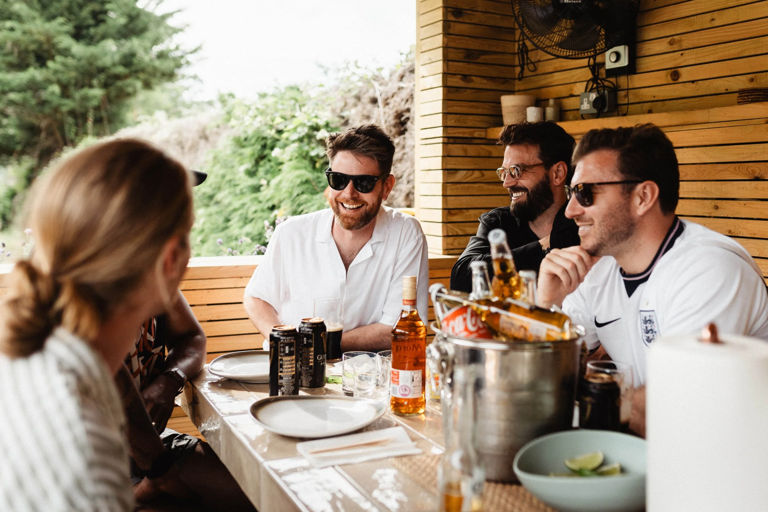 A group of friends is sitting at a table outdoors, enjoying drinks and socializing, with a wooden house and green trees in the background.