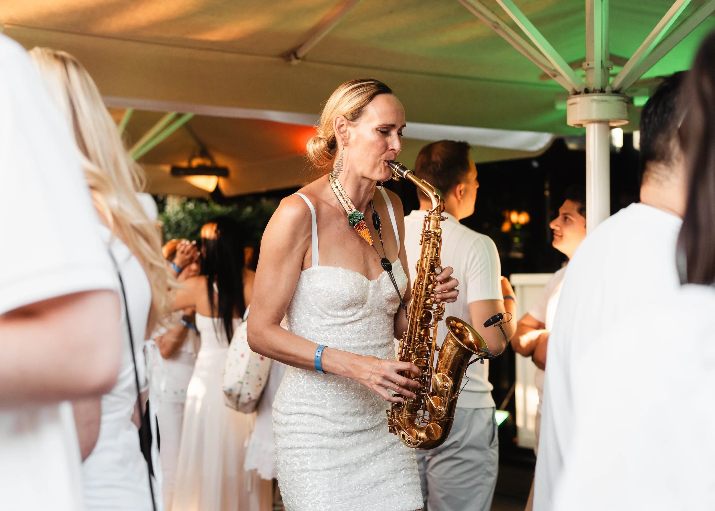 A woman in a white dress playing a saxophone at an outdoor event with other people around her, some of whom are also in white attire.