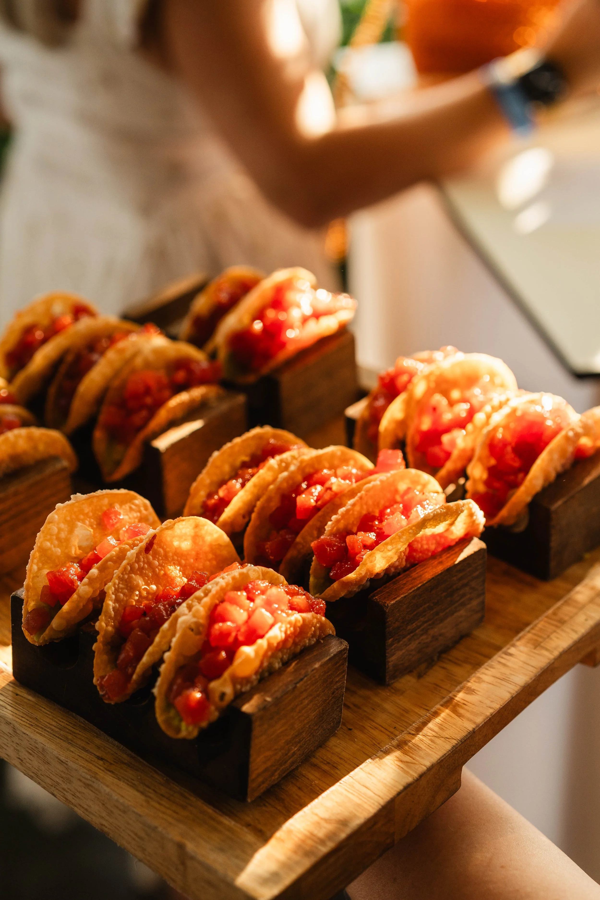 Close-up of a tray of mini tacos filled with diced tomatoes and meat, arranged in wooden compartments on a wooden serving tray.