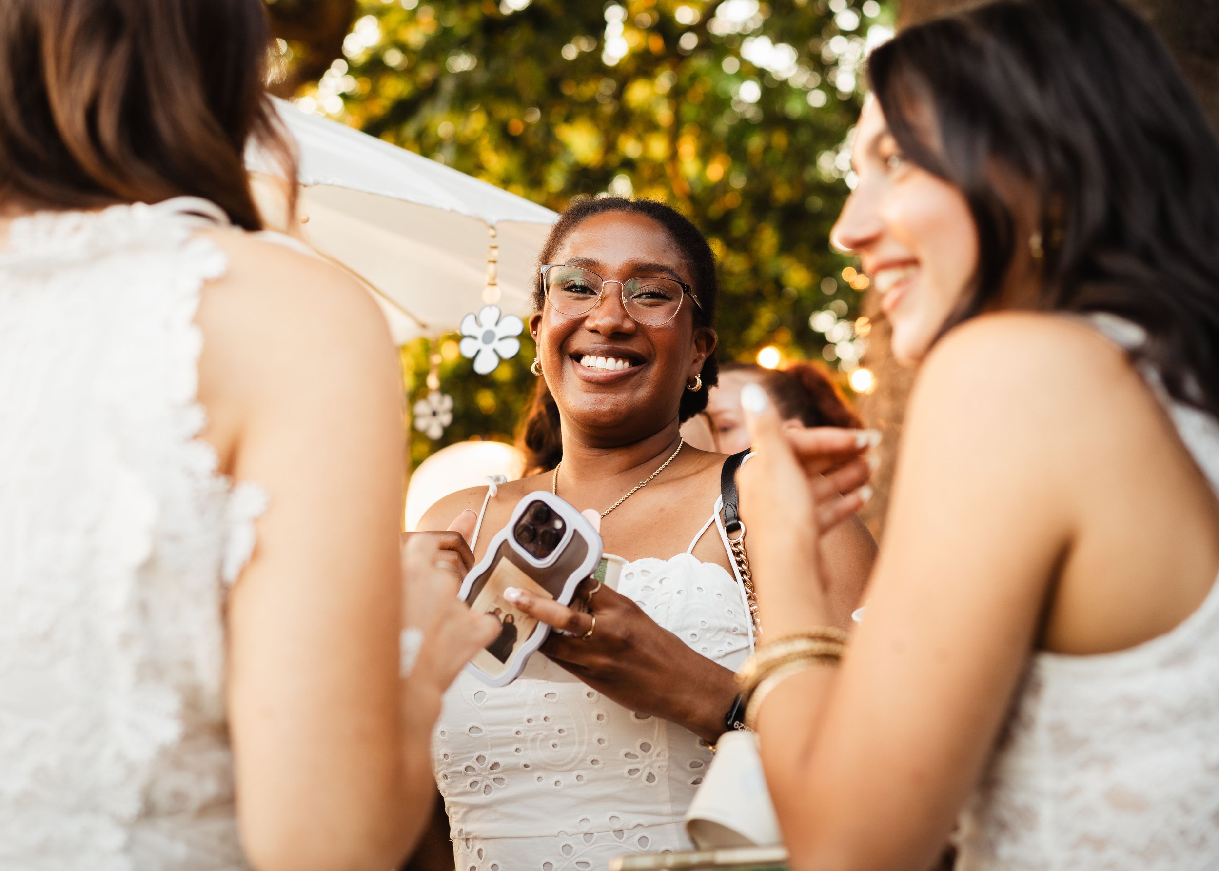 Three women smiling and talking outdoors, one holding a phone, with trees and string lights in the background.