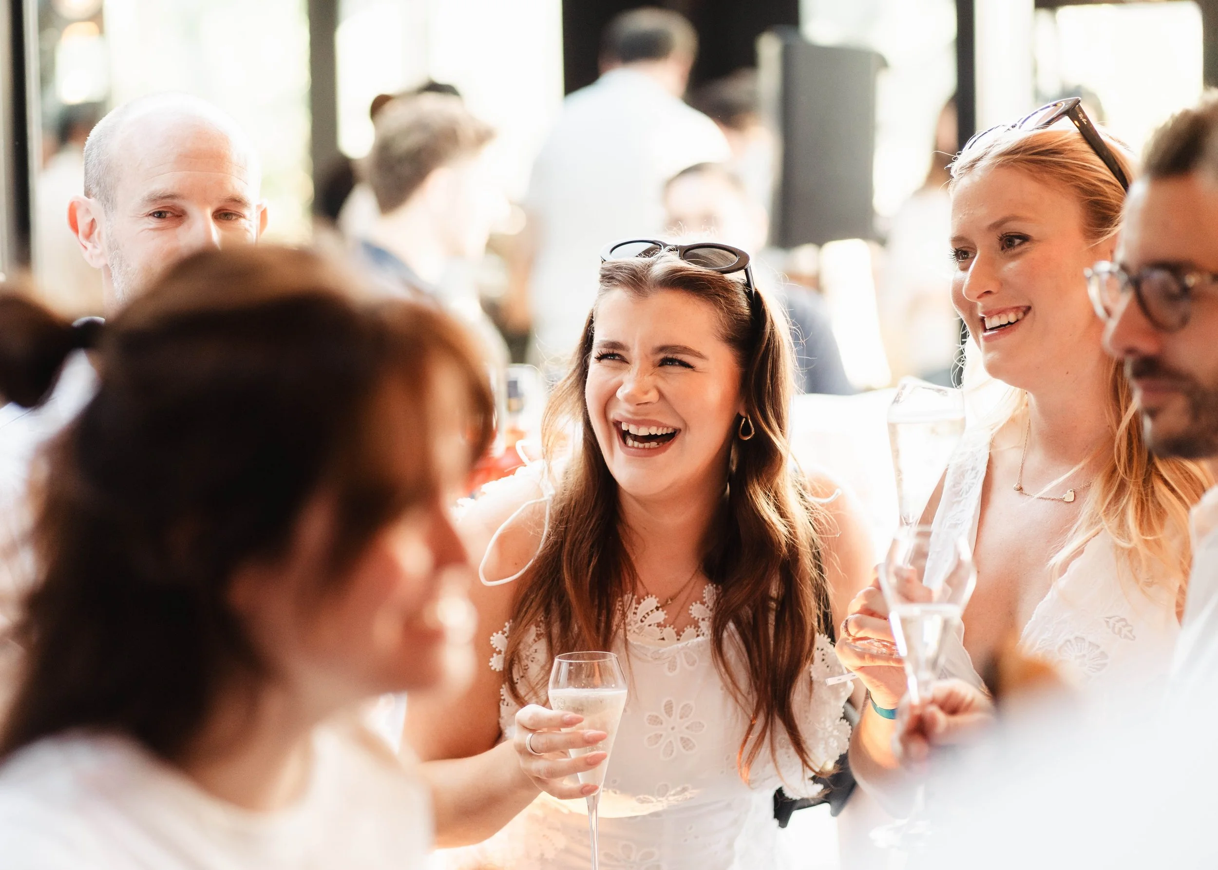 People at a social gathering, mostly women, smiling and holding glasses of champagne or wine, in a bright, crowded indoor setting.