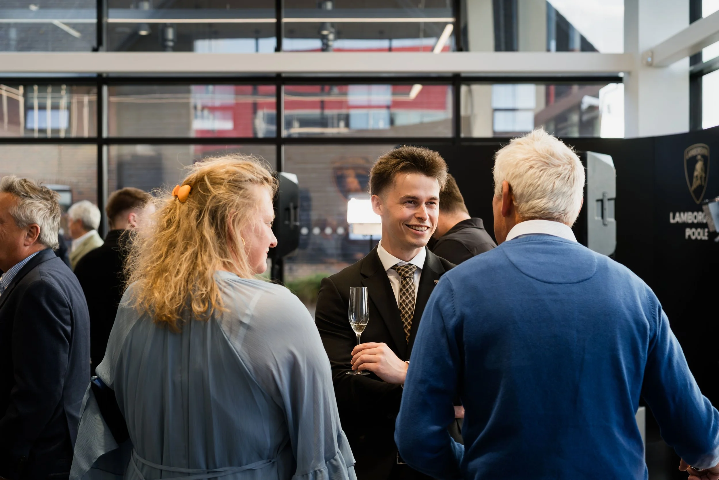 Group of people socializing at a formal event in a modern venue with large windows, including a woman with curly hair and a man with gray hair, and a young man in a suit holding a glass of champagne.