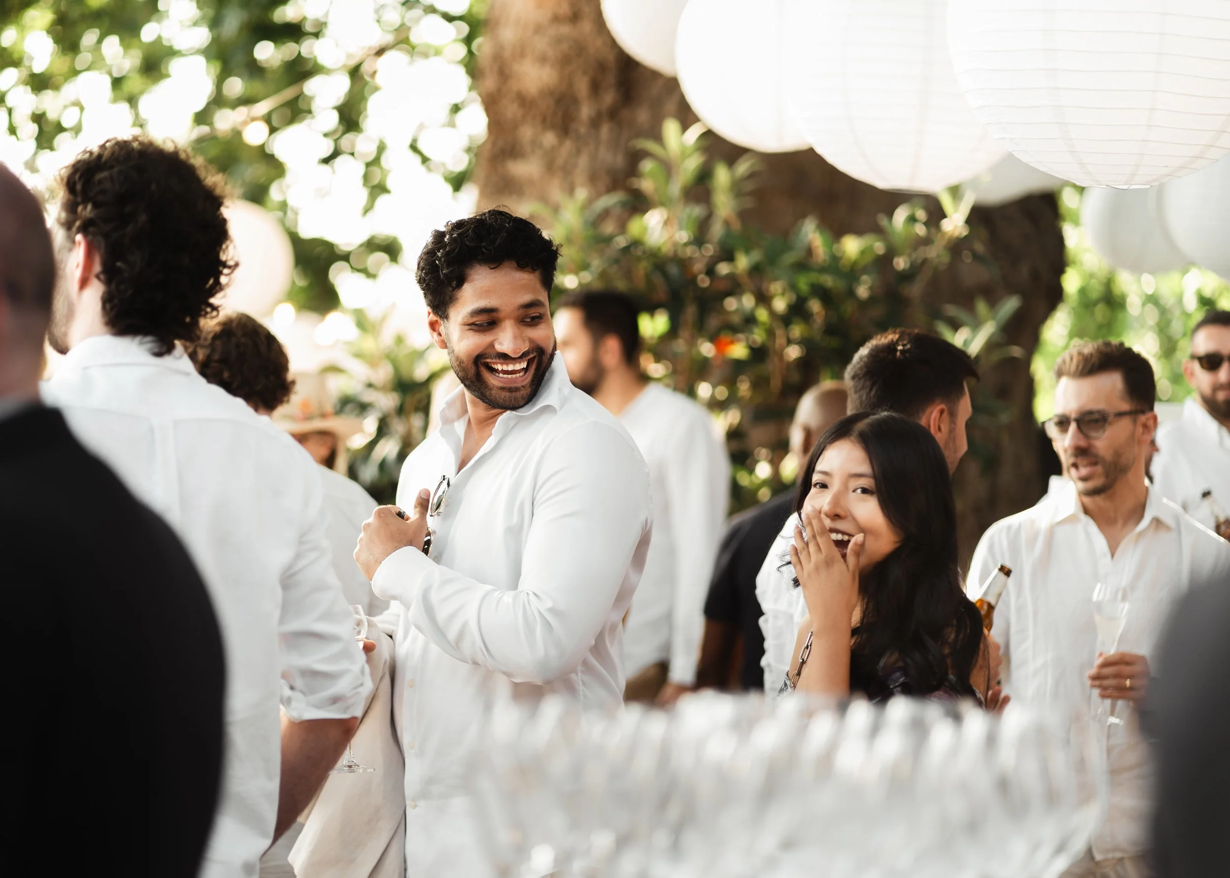 People at an outdoor gathering under hanging white paper lanterns, some holding drinks, socializing, and smiling.