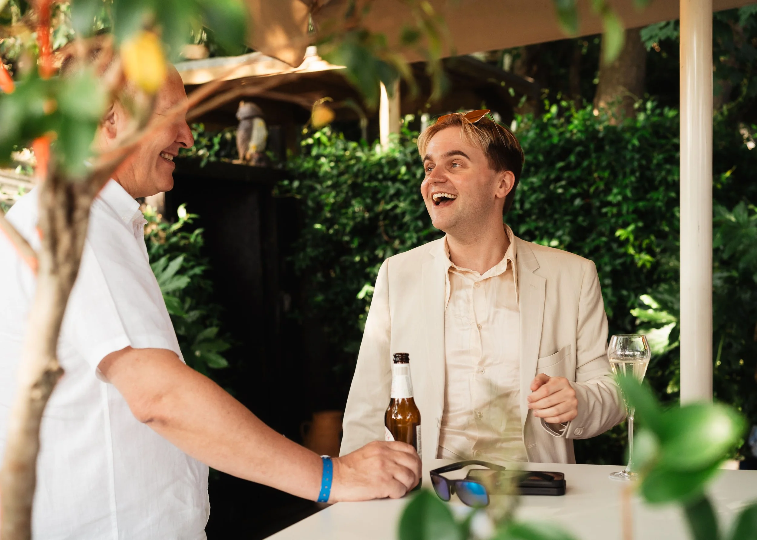 Two men smiling and talking at an outdoor event, one holding a beer bottle, surrounded by greenery and a white table with drinks.