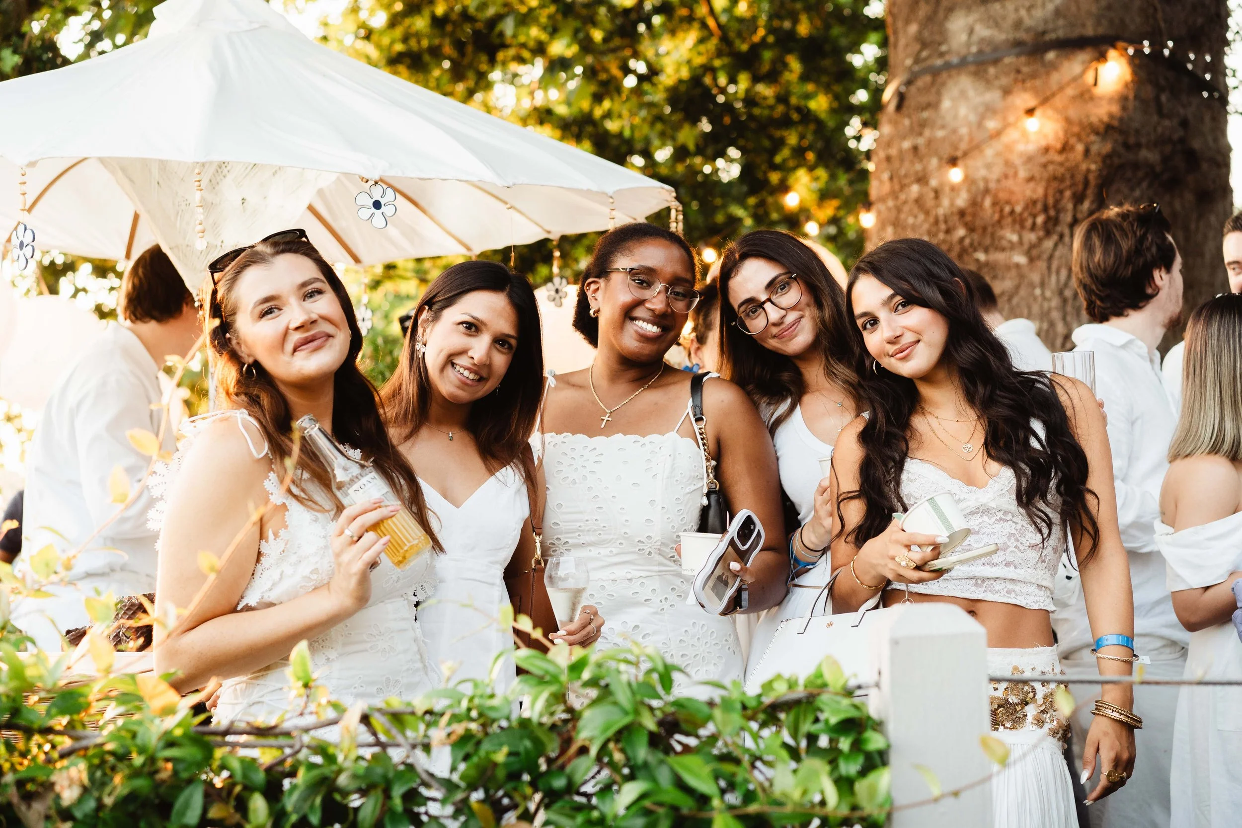 Group of women smiling and posing for a photo at an outdoor event with trees and string lights in the background.