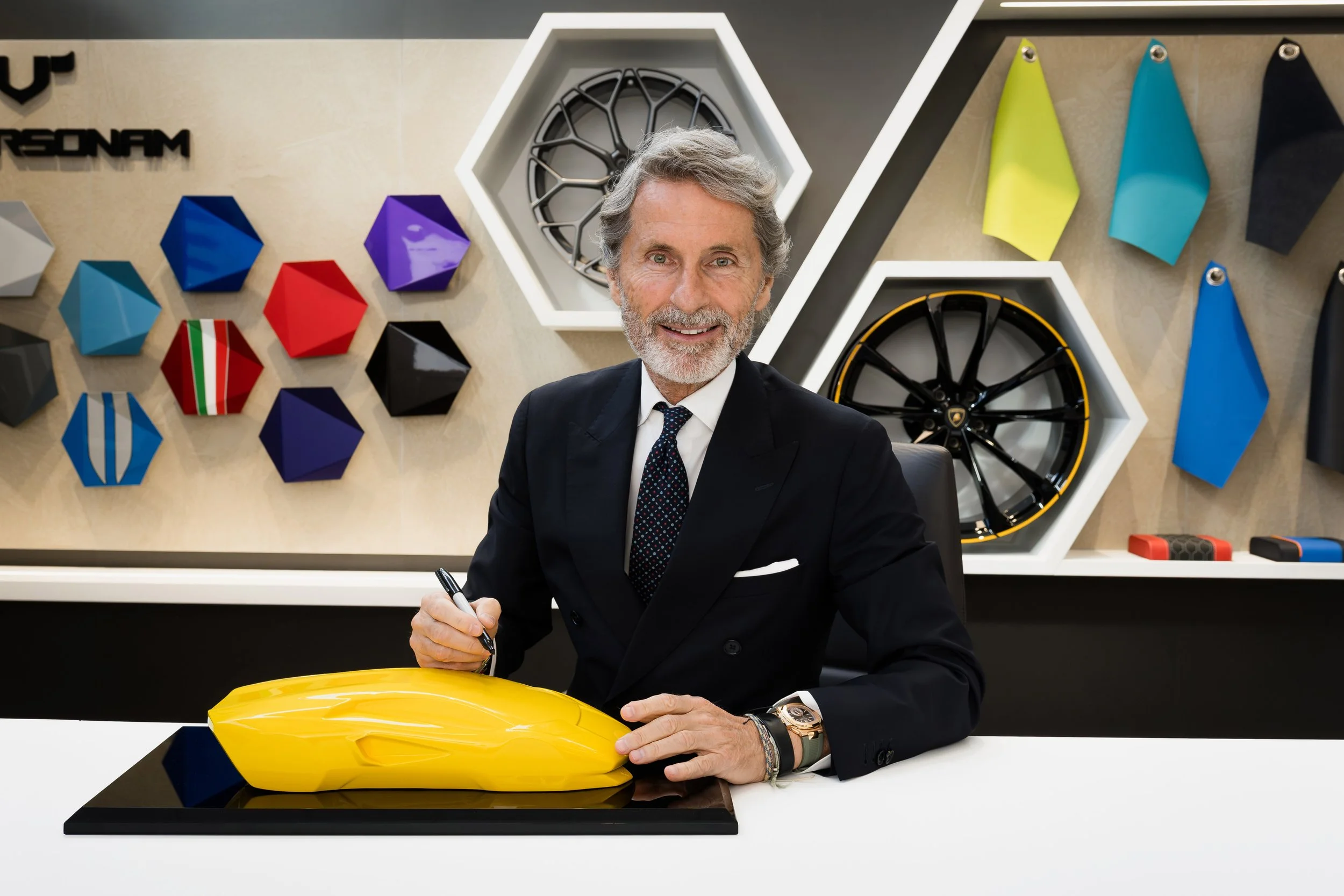 Senior man in suit sitting at a table, holding a pen, next to a yellow car model, in a modern display room with colorful car wheel and panel designs on the wall behind.