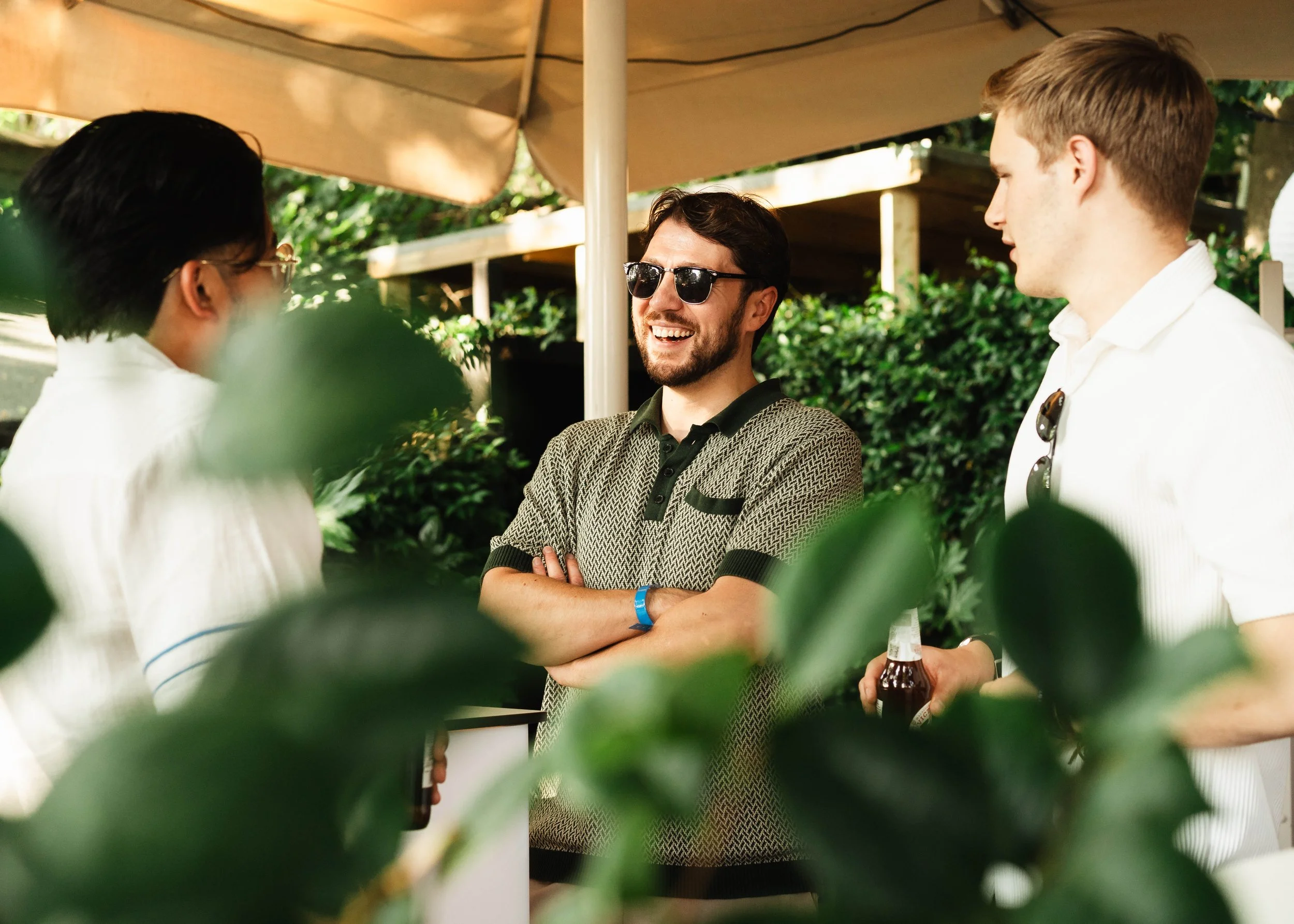 Three men are conversing outdoors under a beige canopy. One is wearing sunglasses and a patterned shirt, smiling with arms crossed. The other two are partially visible, one with glasses and dark hair, and the other with light brown hair, holding a be