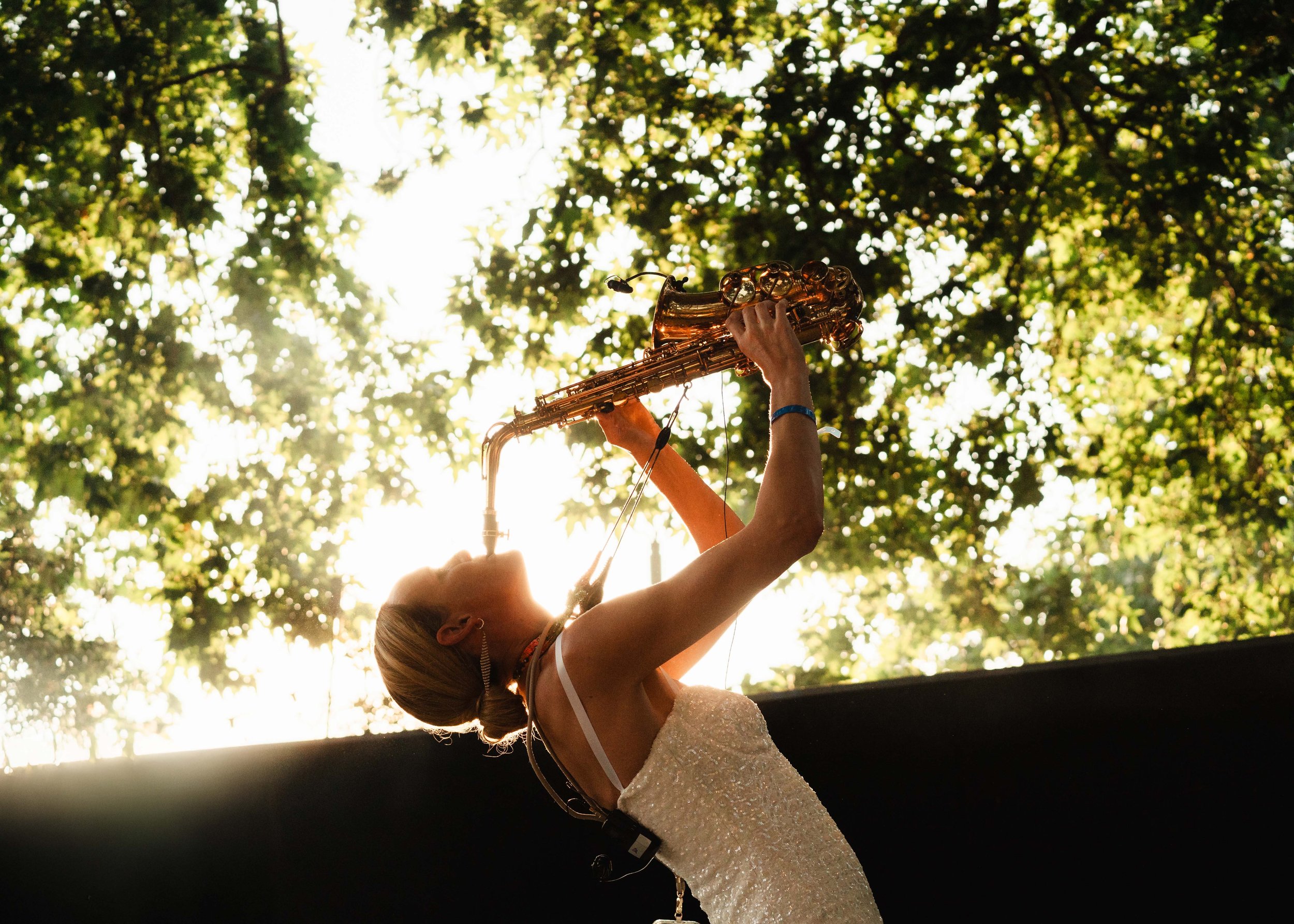 A woman in a white, strapless dress holding a saxophone up towards the sky during daylight, with trees and sunlight filtering through the leaves in the background.