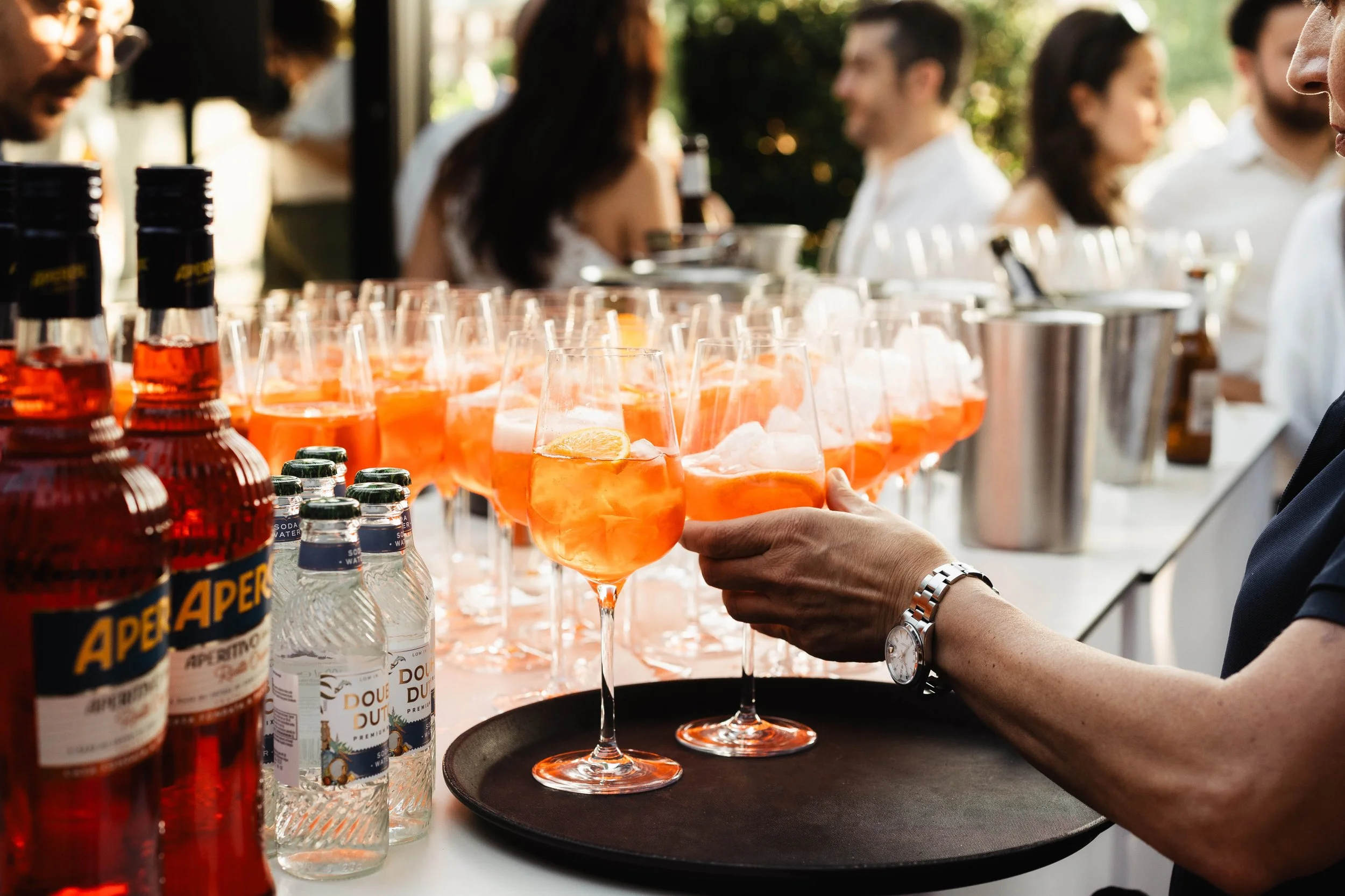 A server is placing glasses of orange drink with lemon slices on a tray at an outdoor event. In the background, several people are socializing, and bottles of beverages are lined up on the table.