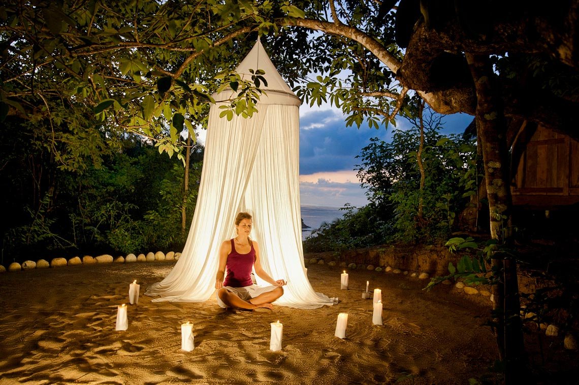 A woman practicing meditation and yoga outdoors at sunset, sitting cross-legged on the ground with a draped fabric canopy behind her, surrounded by lit candles in a natural, tropical setting with trees and ocean in the background.