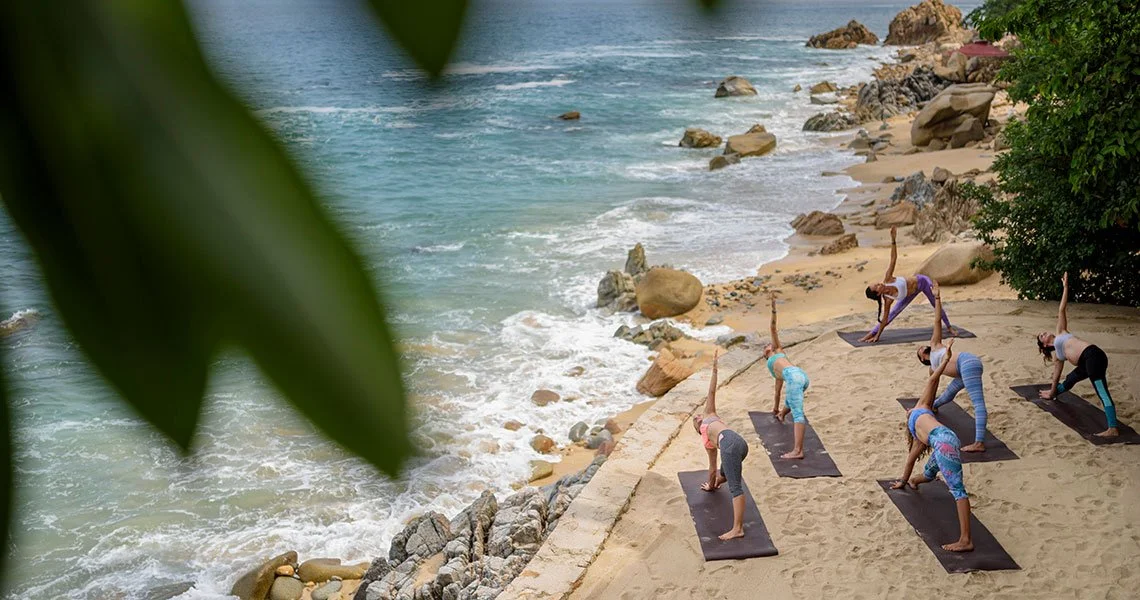 Group of people practicing yoga on beach during sunset, with rocks and ocean in background