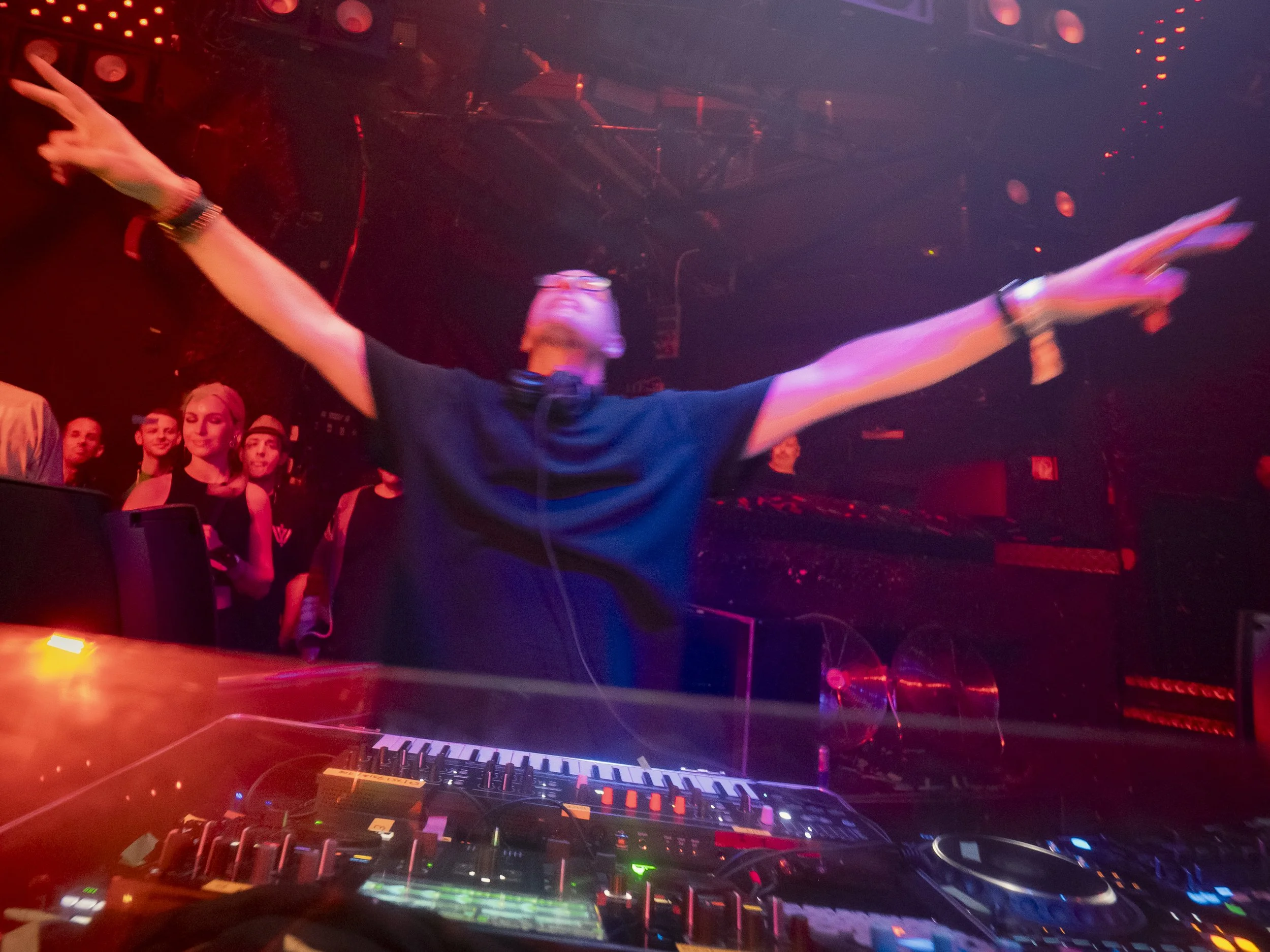 A DJ is performing at a nightclub with arms outstretched, wearing glasses and a headset, surrounded by a crowd of people under dim, colorful lighting.