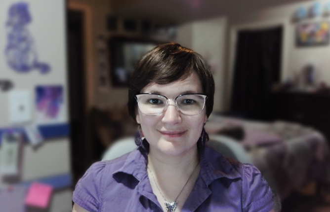 Image is of Carissa, a white woman with short, brown hair and brown eyes. She has large, cat eye glasses and is wearing a dressy purple shirt, dangly purple earrings, and a silver necklace with a purple butterfly. She is smiling at the camera.