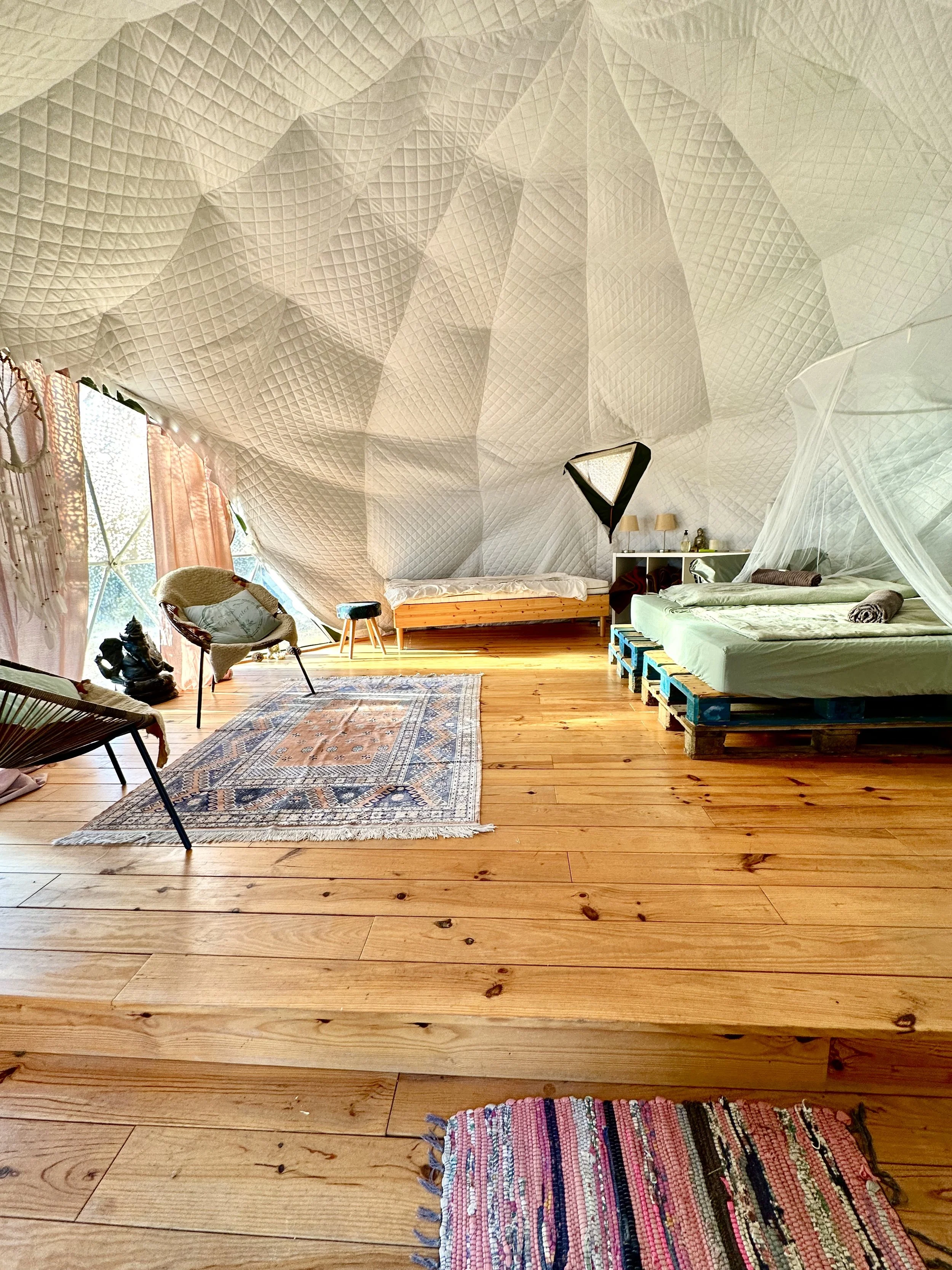 A cozy room with a wooden floor and unique, textured ceiling. It features a bed made from pallets with a mosquito net, a small colorful rug, a vintage-style rug, and seating including a chair and a lounge chair. There's a window with pink curtains allowing natural light, and various decorative items on a shelf.