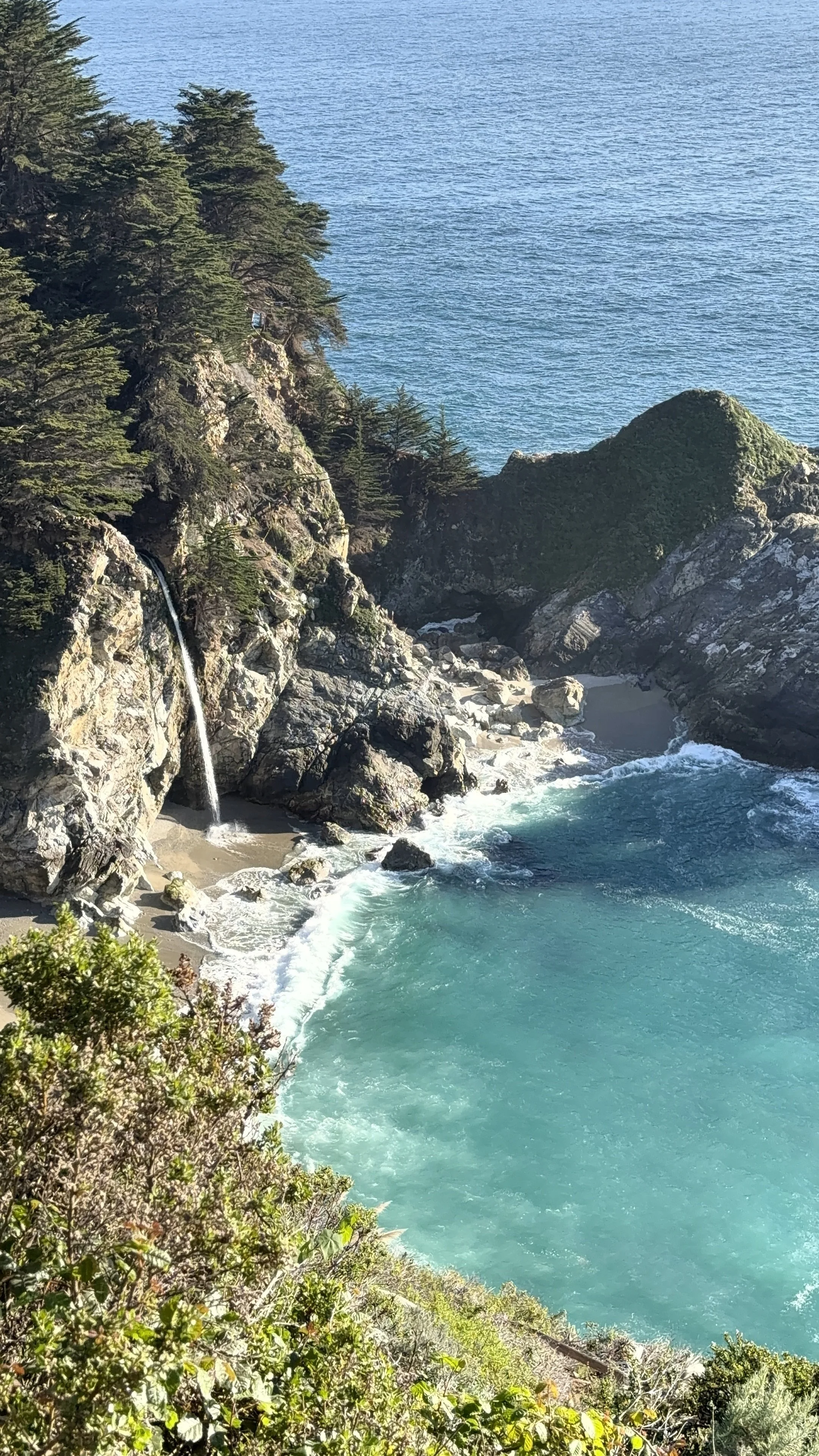 Coastal landscape with a small waterfall flowing from rocks into the ocean, surrounded by green trees and bushes.