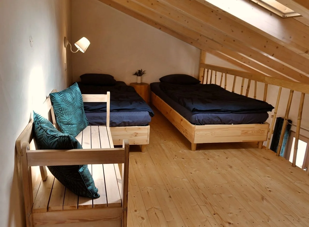 Cozy wooden interior with two beds covered in patterned bedspreads, a small table with a lamp and a fan, a gray sofa, a clothing rack, and an open kitchen in the background.