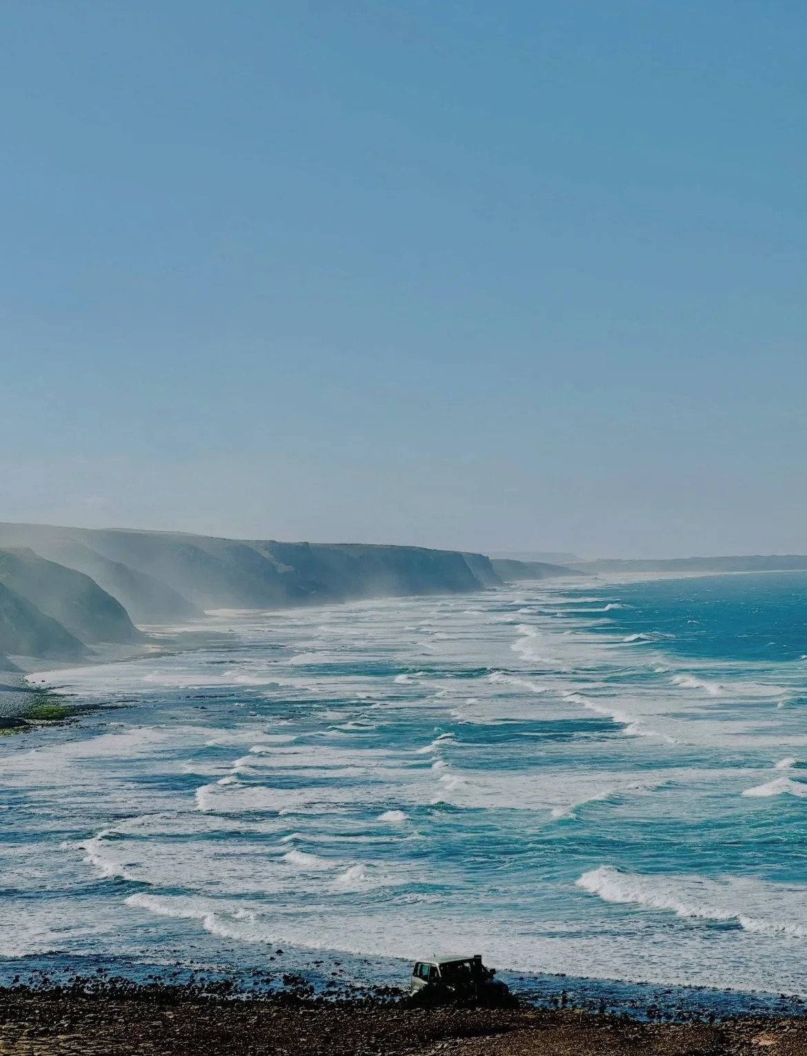 Ocean waves crashing against the rocky shore with cliffs in the background under a clear blue sky, and a vehicle parked on the beach in the foreground.