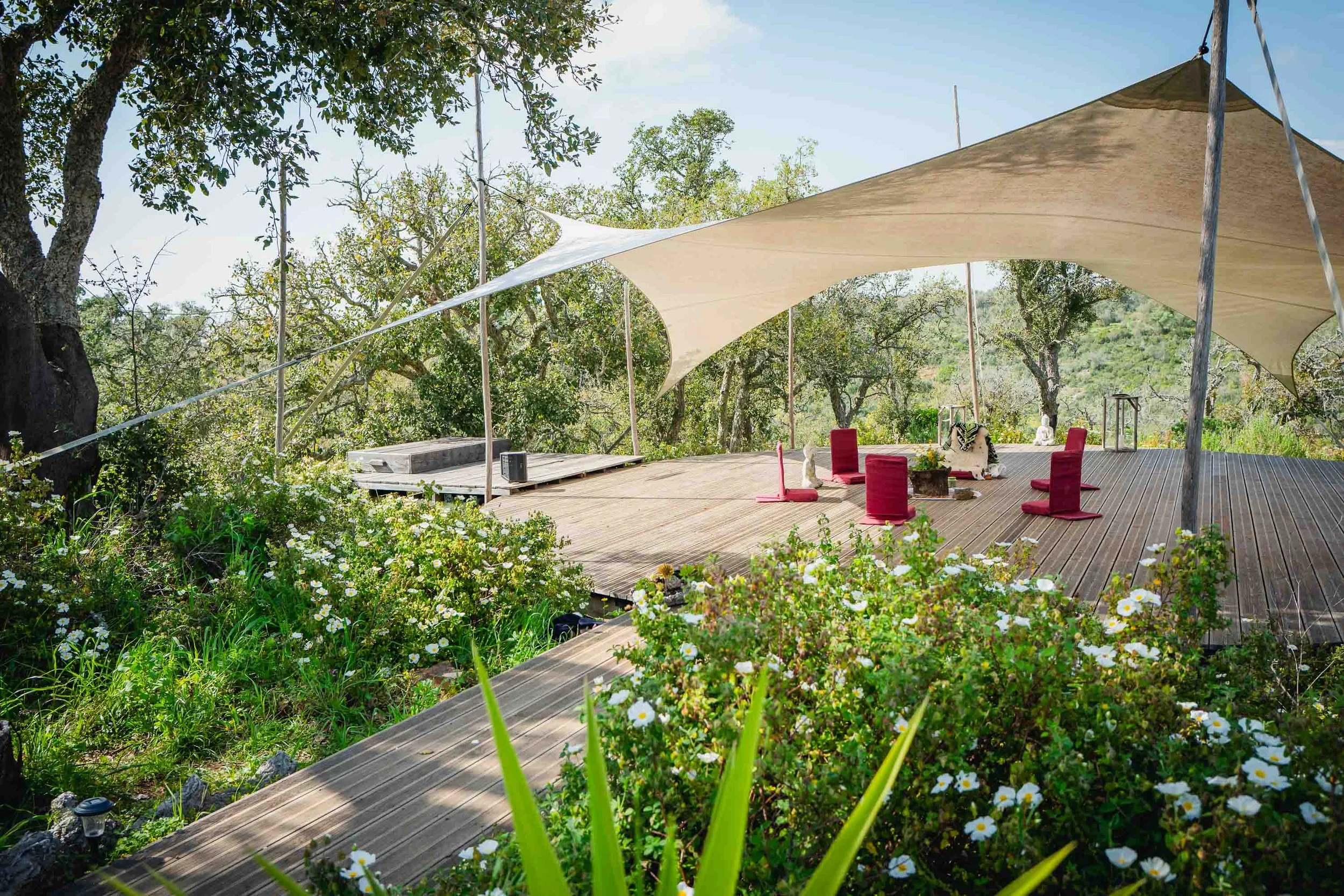 An outdoor wooden deck with a large beige canopy, surrounded by greenery and trees, featuring four red chairs, space for seating, and decorative sculptures.