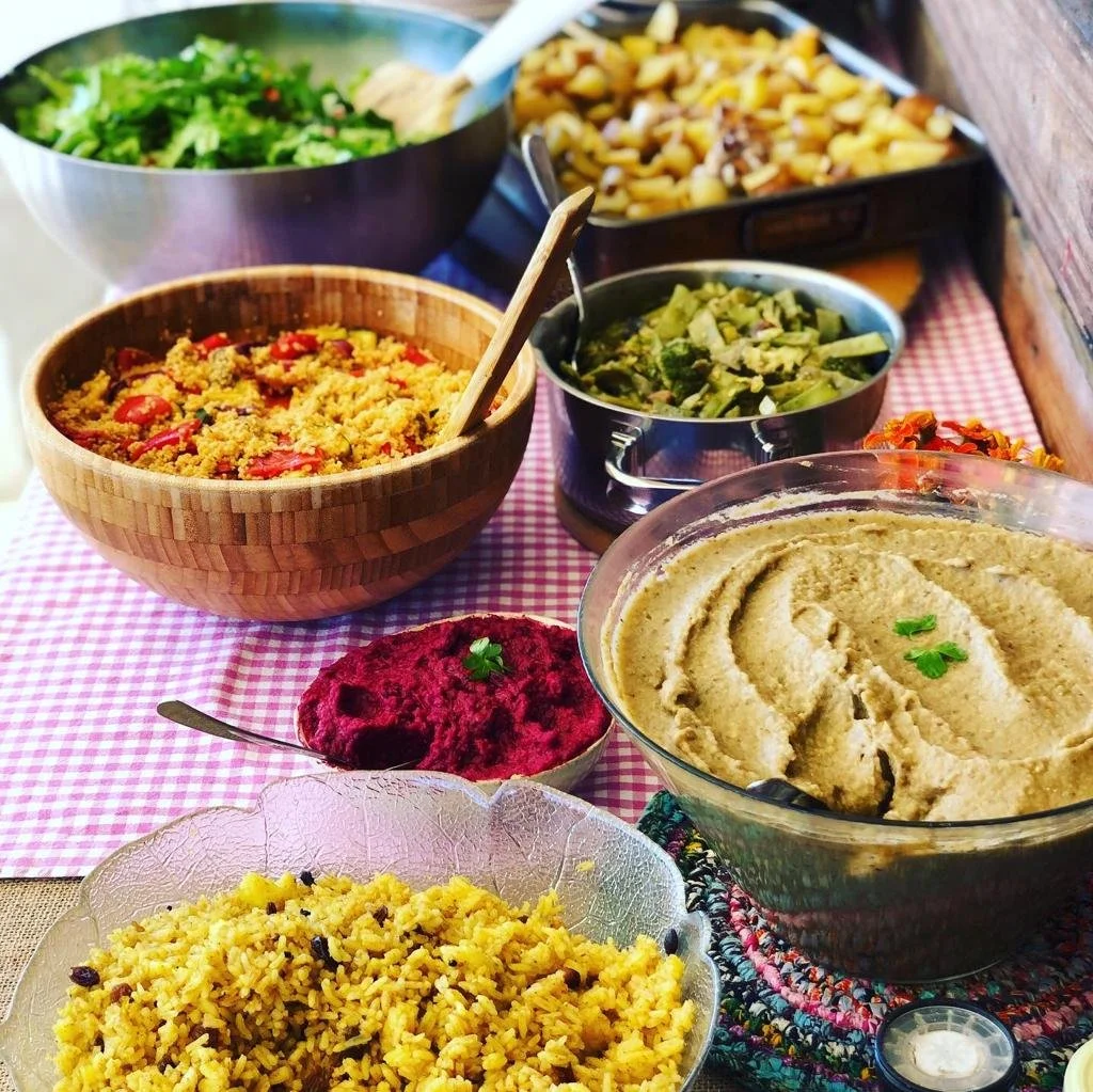 A variety of colorful dishes including salad, rice, hummus, beet dip, and other Mediterranean dishes on a table with a pink checkered tablecloth.
