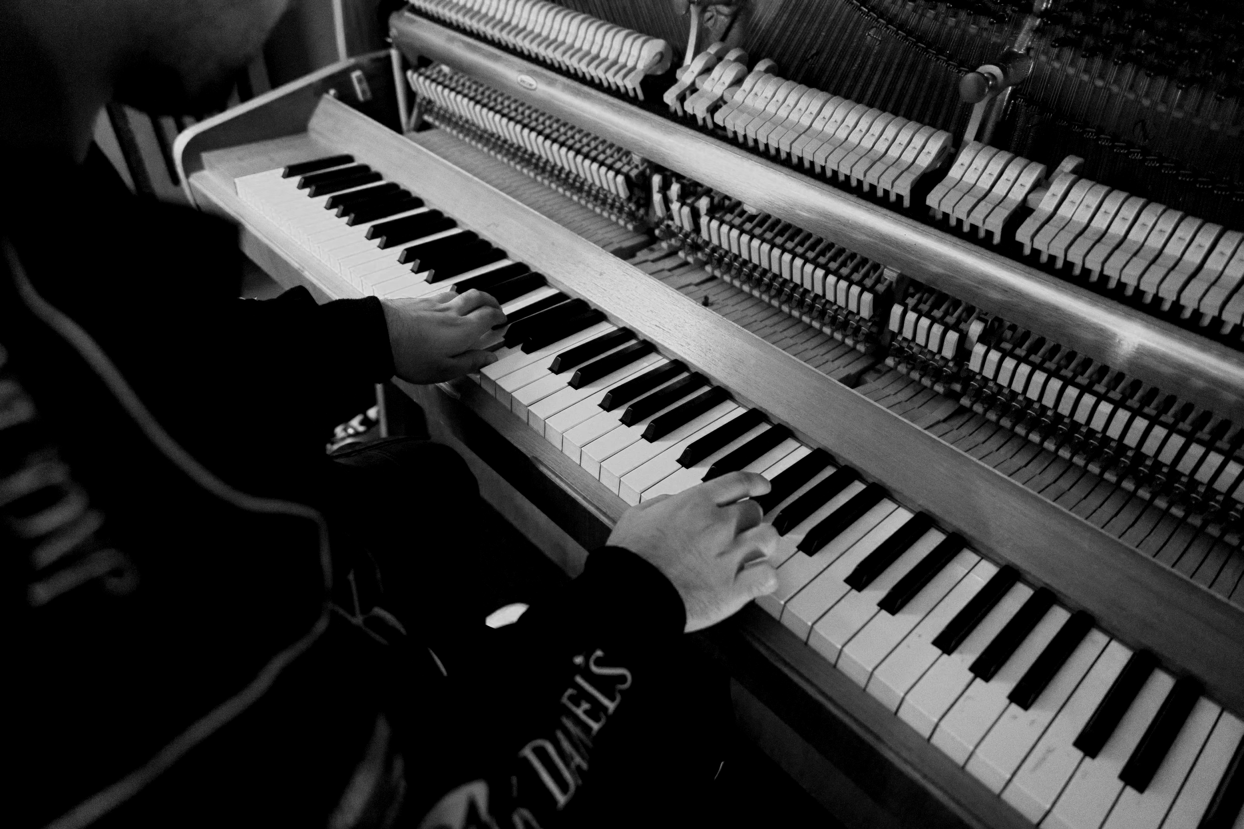 A person playing an upright piano, with focus on their hands on the keyboard, in black and white.