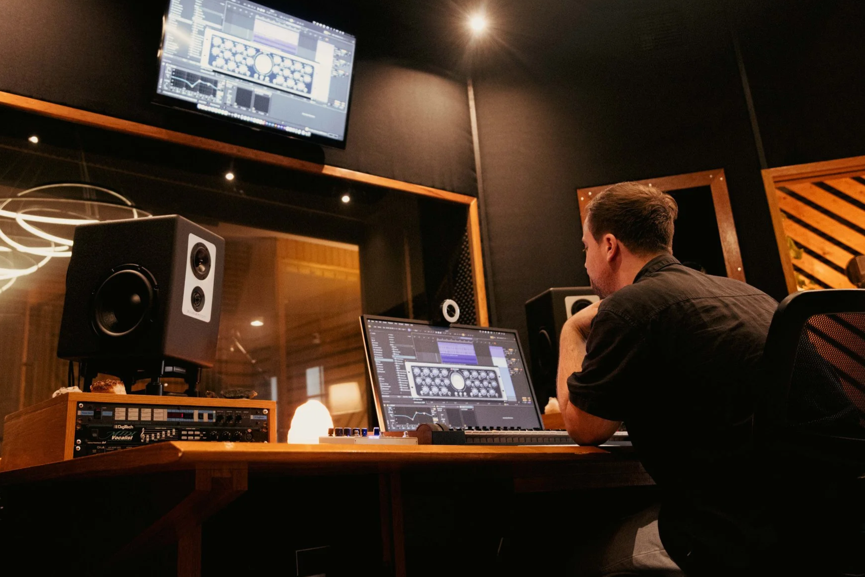 A person working in a music studio with digital audio workstation on laptop and monitor, studio speakers on desk, and soundproof room.