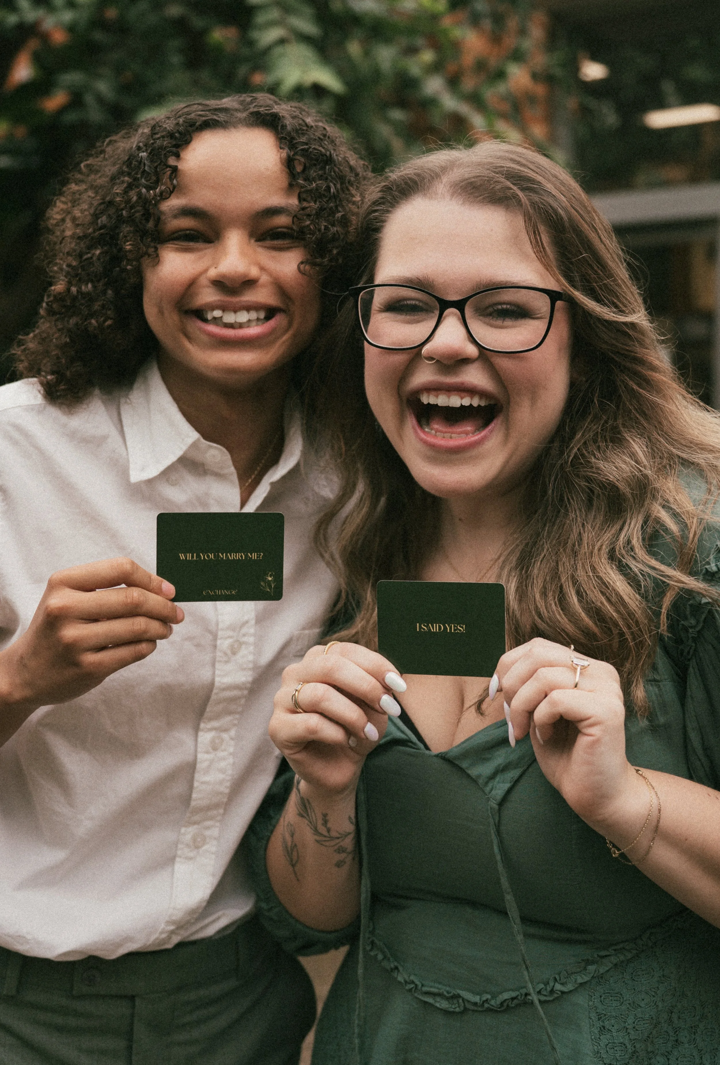 two recently engaged women smiling and  and holding up cards that say "will you marry me?" and "I said yes!"