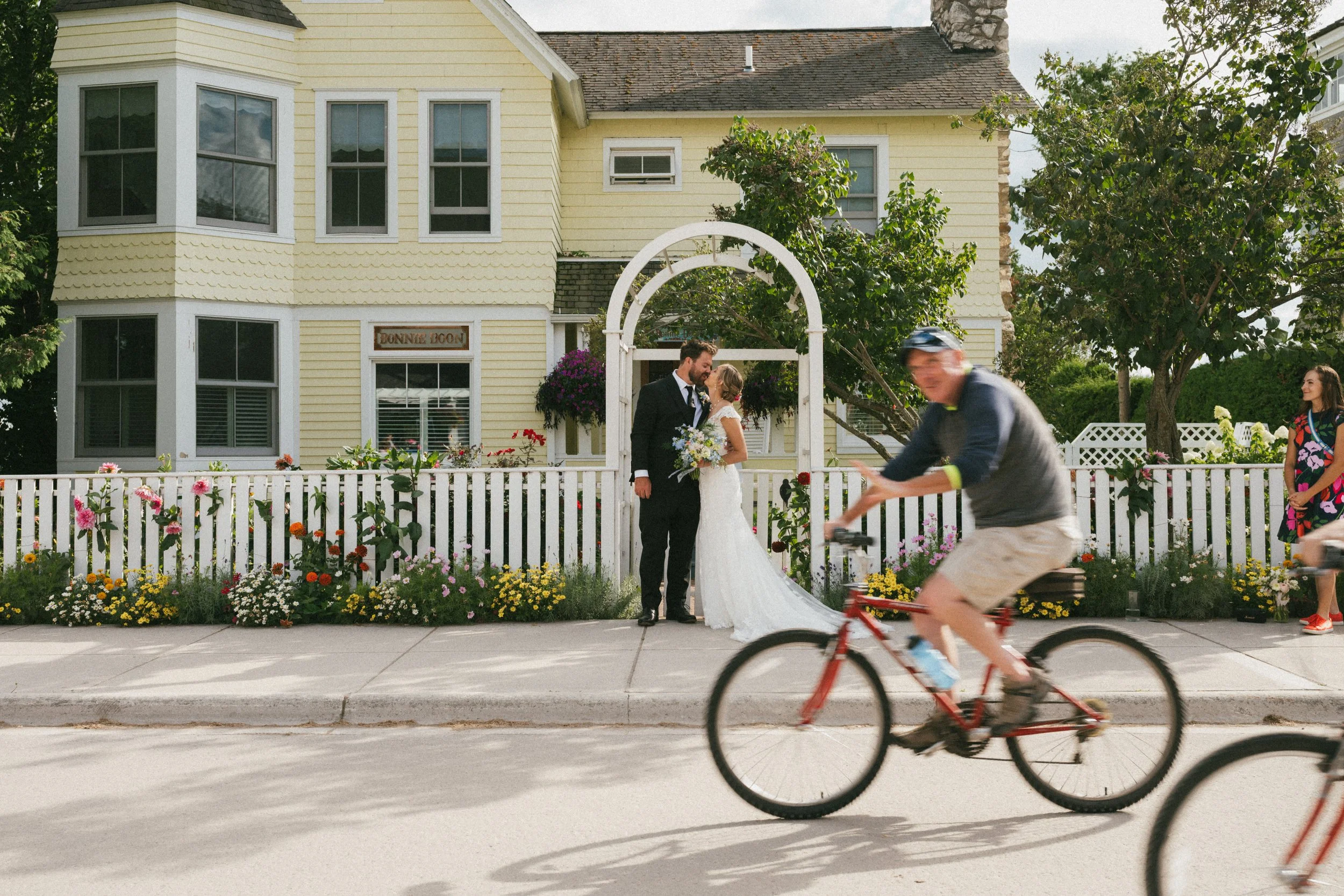 bride and groom kissing in front of a yellow house while bikers whizz by in the foreground