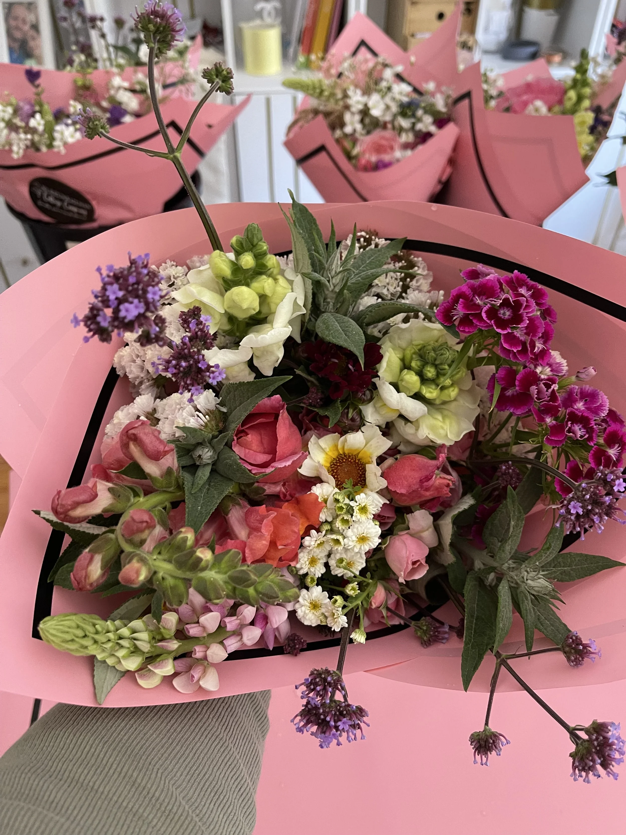 A vibrant bouquet of mixed flowers in a pink wrapping, featuring roses, daisies, and snapdragons with varied greenery and small purple flowers, arranged on a table in a floral shop.