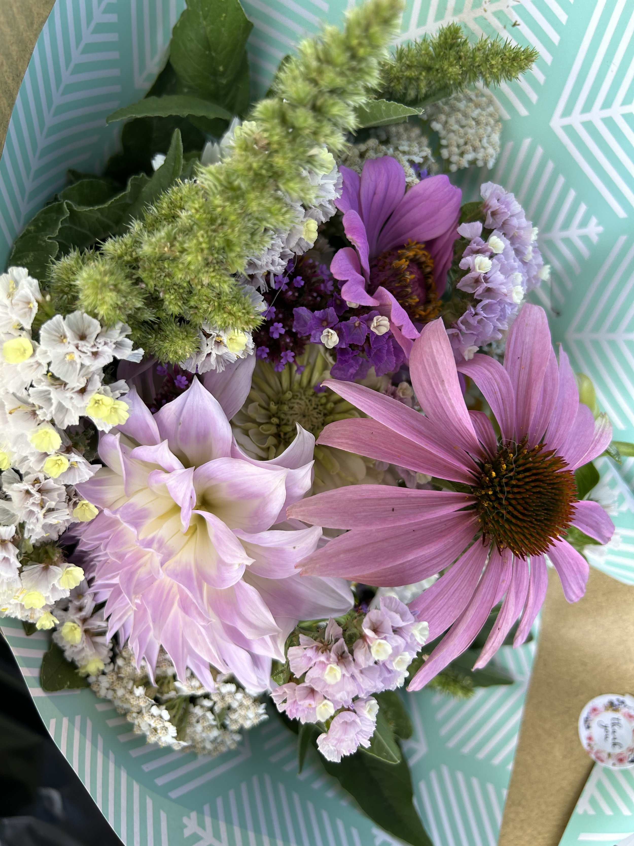 A bouquet of mixed flowers featuring a pink Echinacea, a pale purple dahlia, and small pastel blossoms, arranged on a light teal patterned background with zigzag lines.